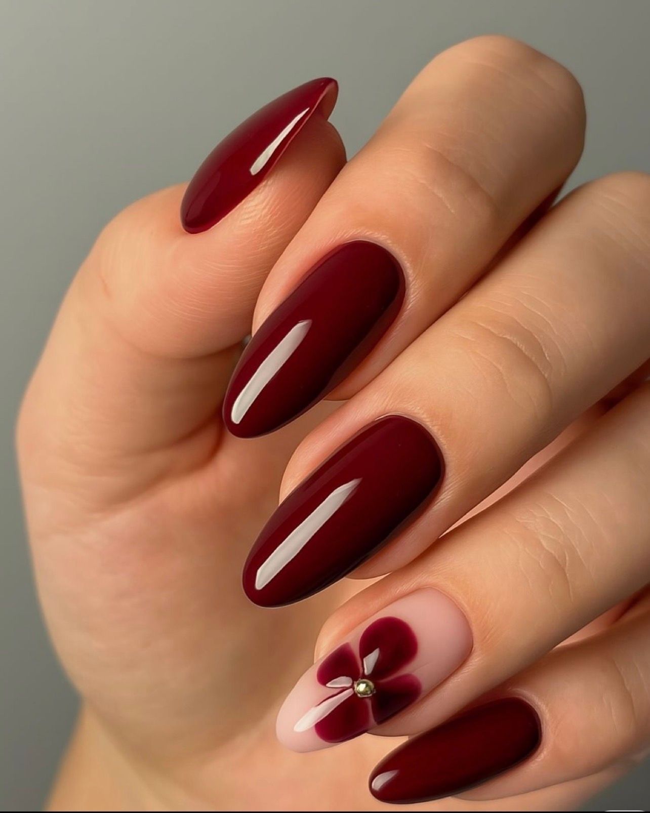 Main aux ongles longs en amande de couleur bordeaux ; un ongle présente un motif floral rose et bordeaux avec un centre en pierre précieuse.