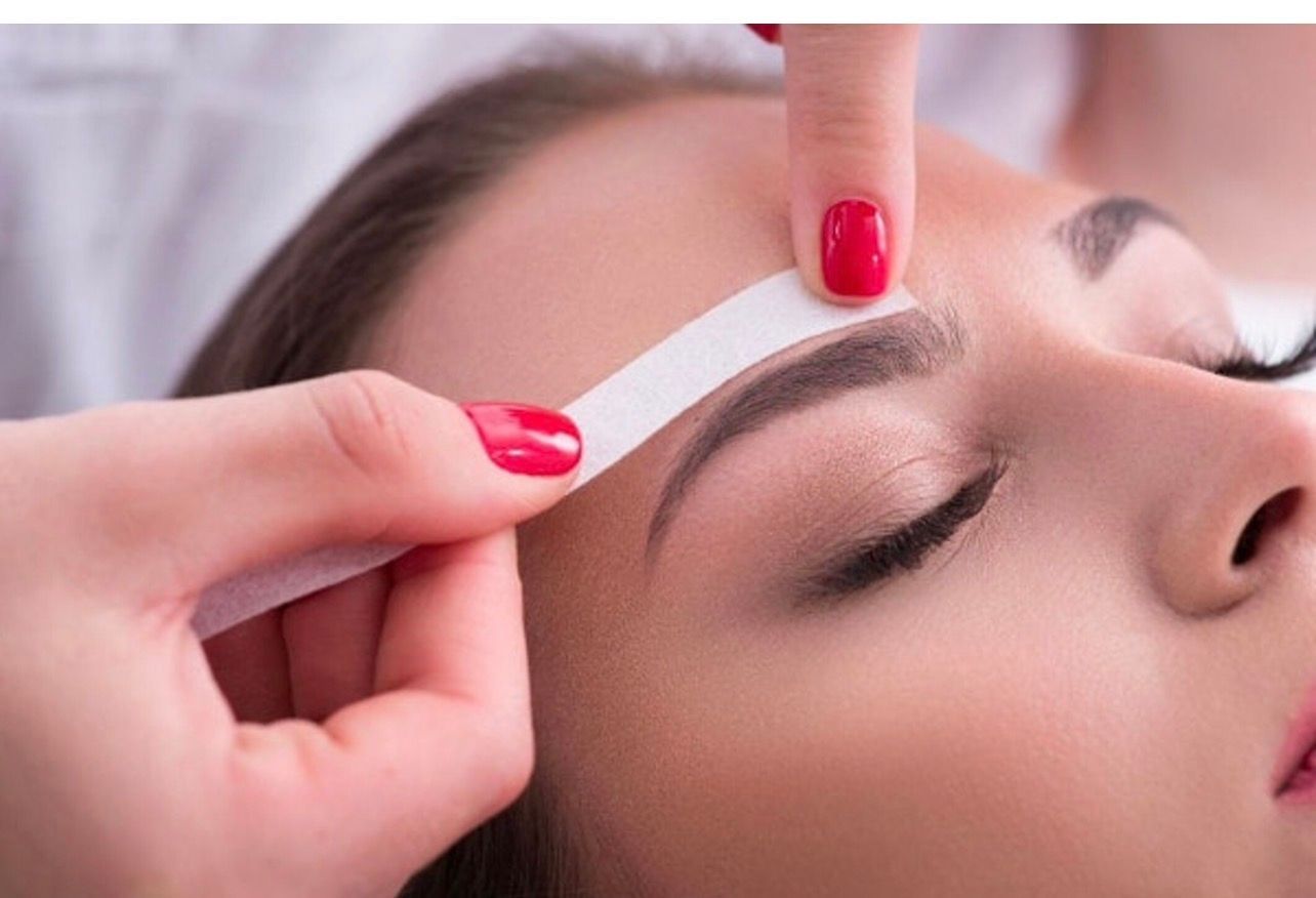 Une femme se fait épiler les sourcils à la cire ; quelqu'un lui applique une bandelette de vernis à ongles rouge sur le sourcil.