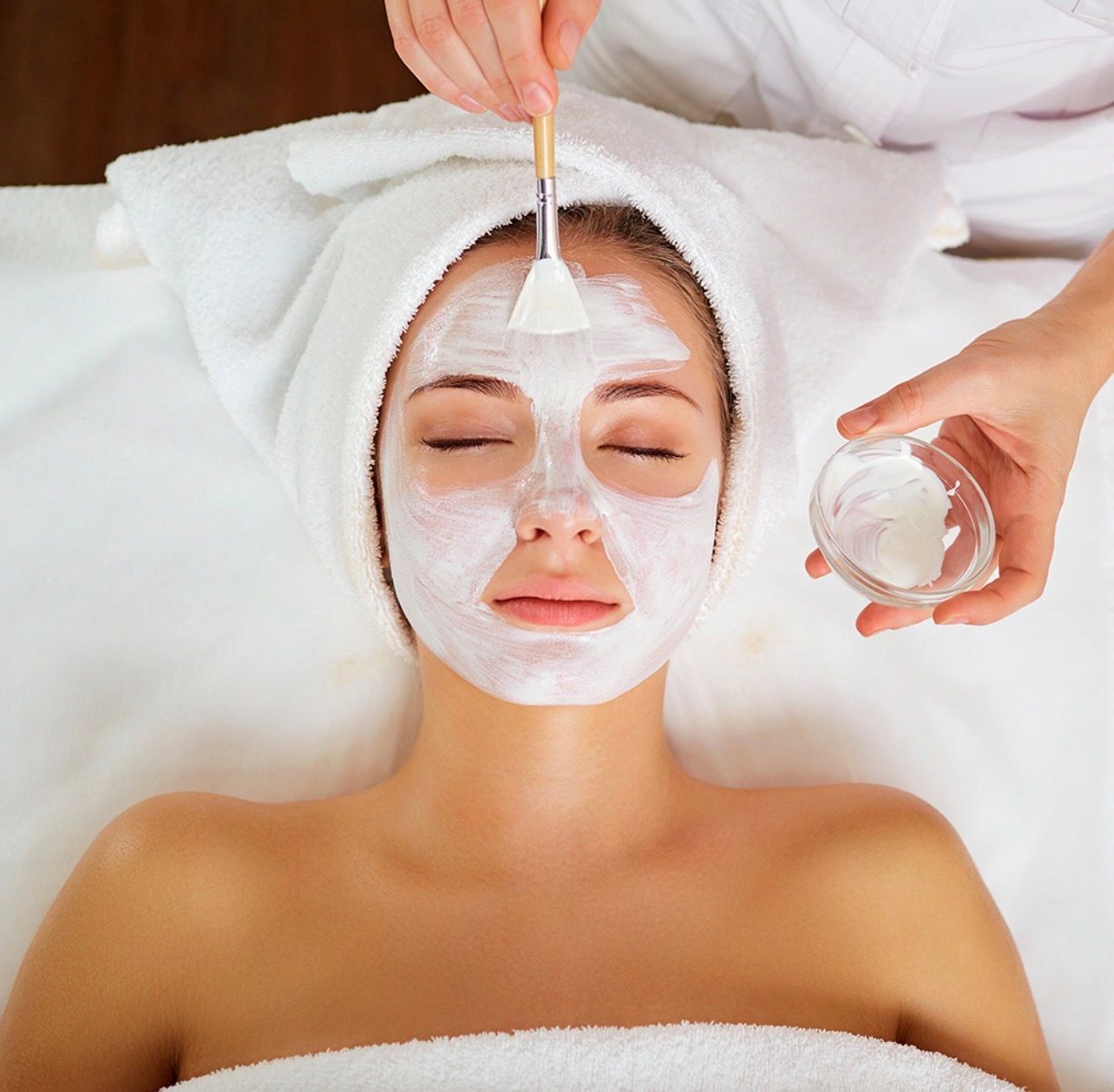 Une personne reçoit un masque facial dans un spa, les yeux fermés, une serviette blanche sur la tête, les mains appliquant le masque.