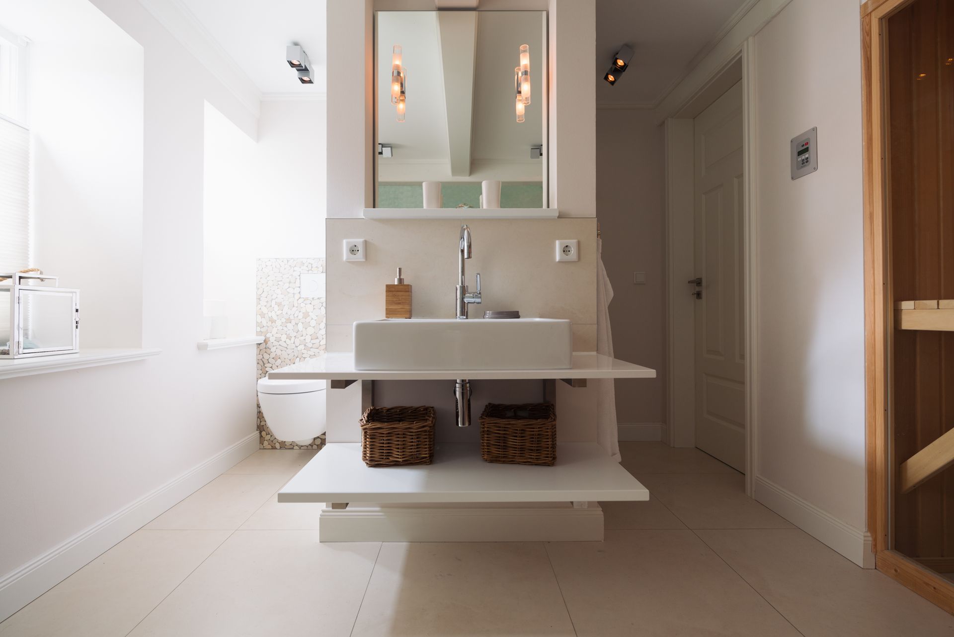 Salle de bains d’un blanc éclatant avec lavabo sur pied, armoire de toilette avec miroir, toilettes et paniers en osier sur une étagère.