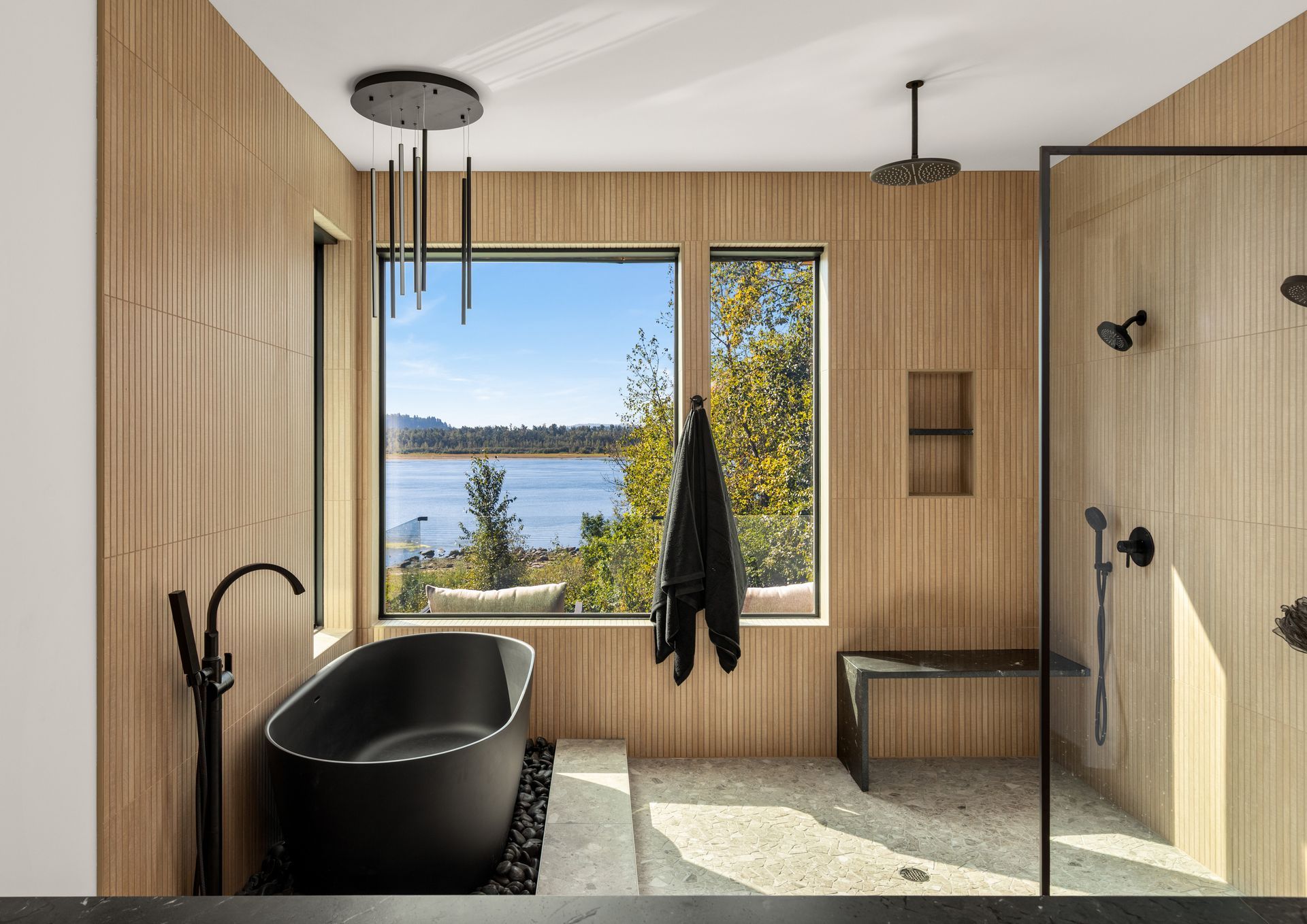 Salle de bains moderne en bois avec baignoire noire, douche et vue sur un lac à travers une grande fenêtre.