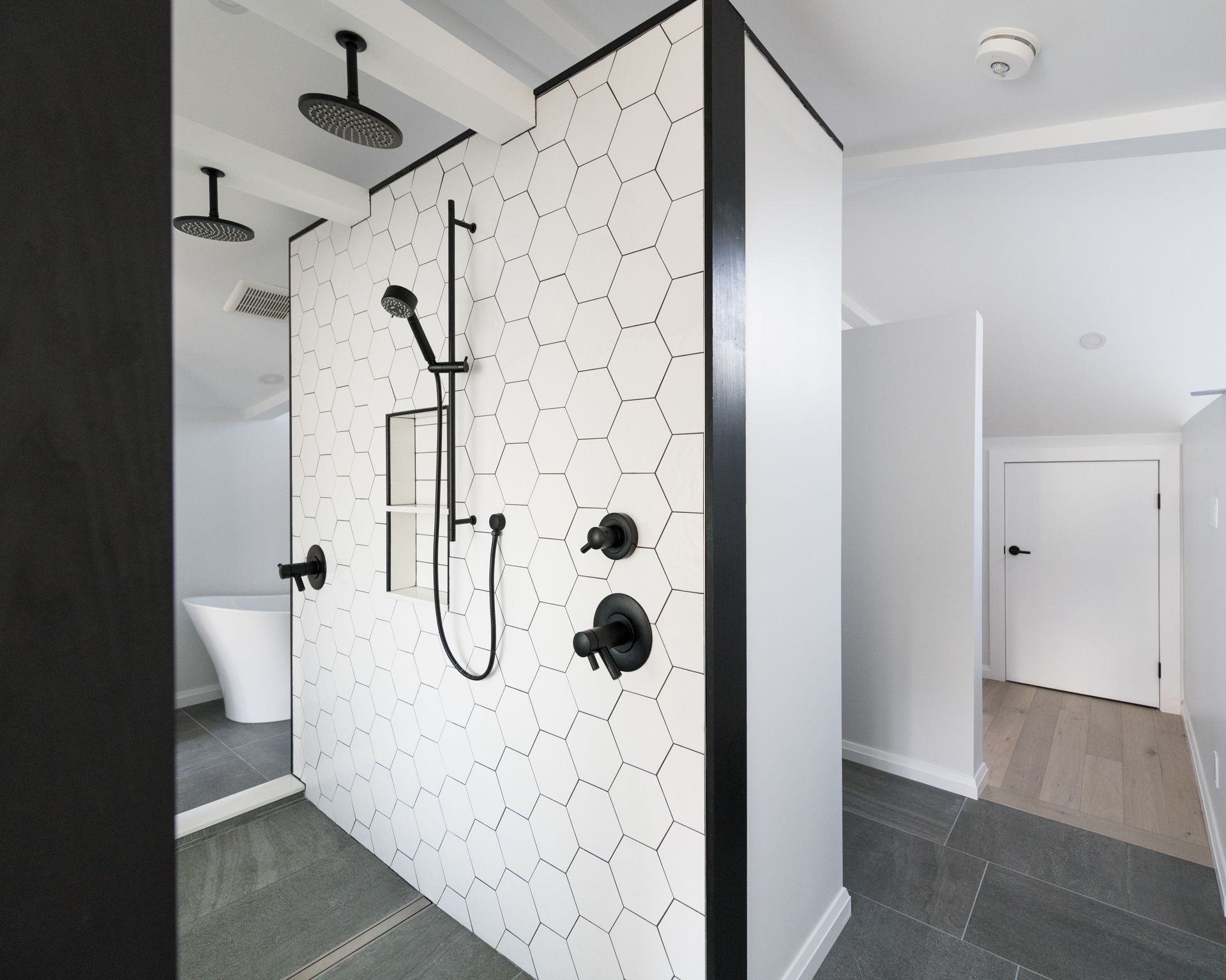 Douche moderne dans une salle de bains avec du carrelage hexagonal blanc, des robinetteries noires et une entrée de couloir attenante.