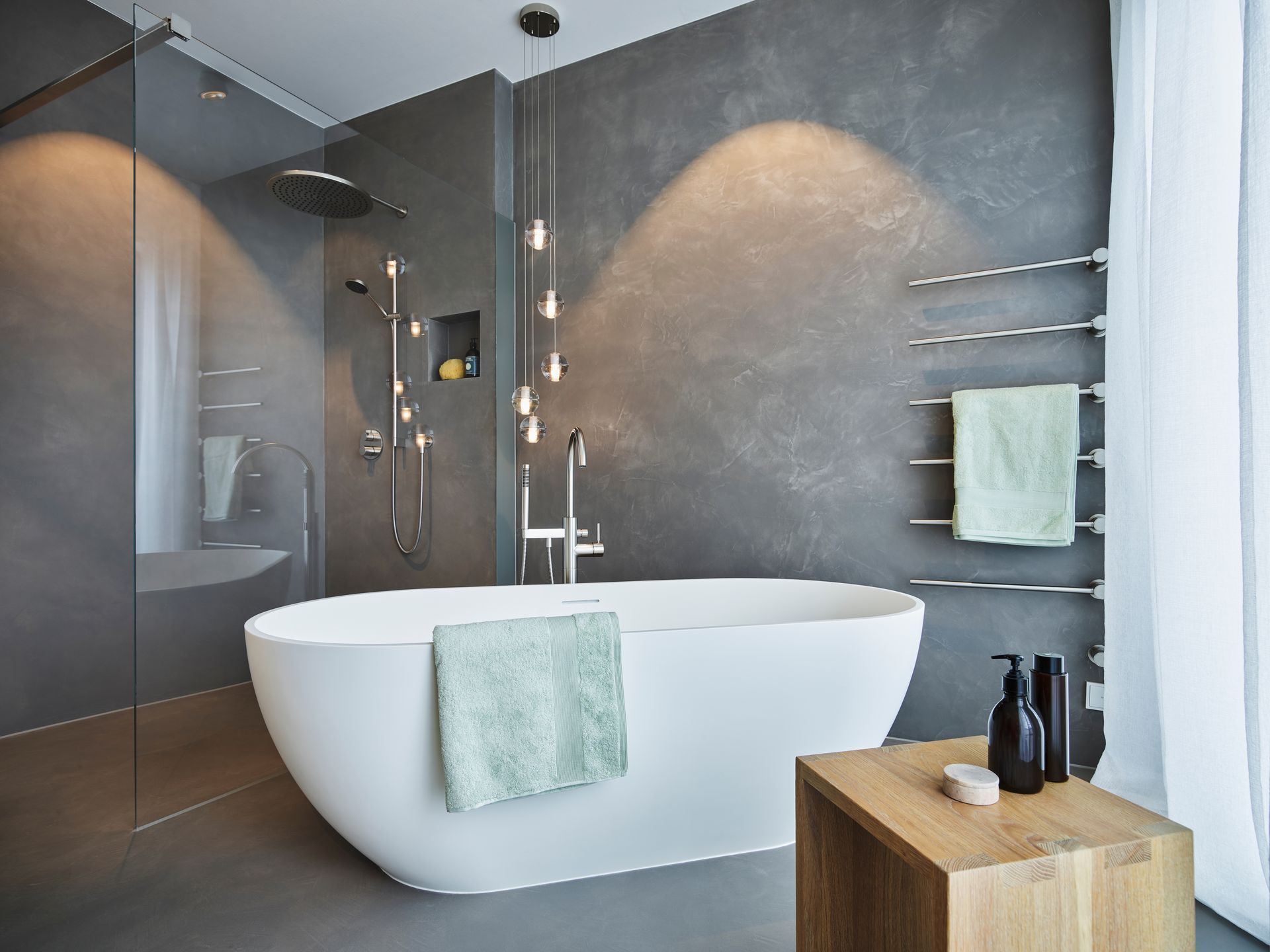 Salle de bains moderne avec baignoire blanche îlot, murs carrelés gris, douche vitrée et table d’appoint en bois.