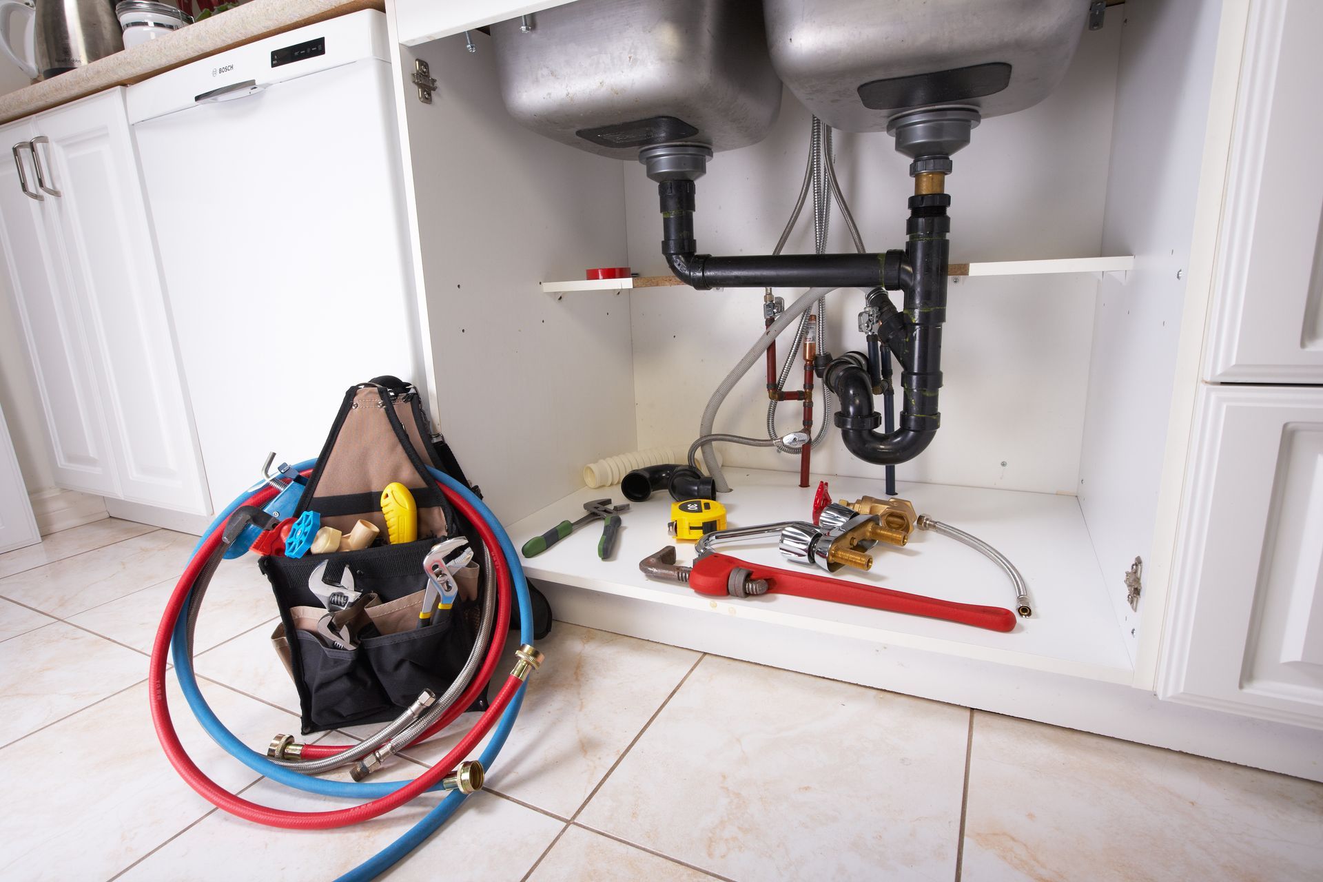 Installation de réparation de plomberie sous évier, avec des tuyaux rouges et bleus, des outils et des conduites apparentes dans un meuble blanc.