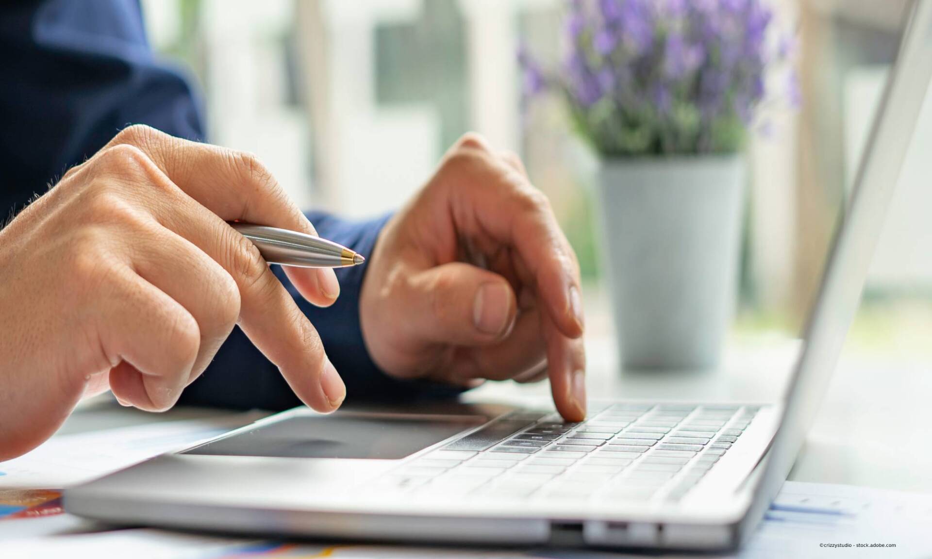 Person tippt mit Stift in der Hand auf Laptop. Nahaufnahme im Innenbereich mit floralem Hintergrund.