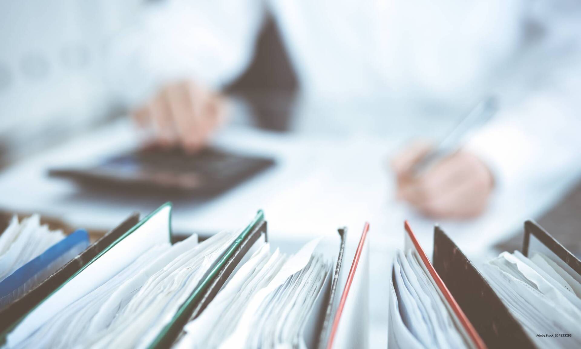 Person, die einen Taschenrechner und einen Stift verwendet, im Vordergrund ein Stapel Akten.