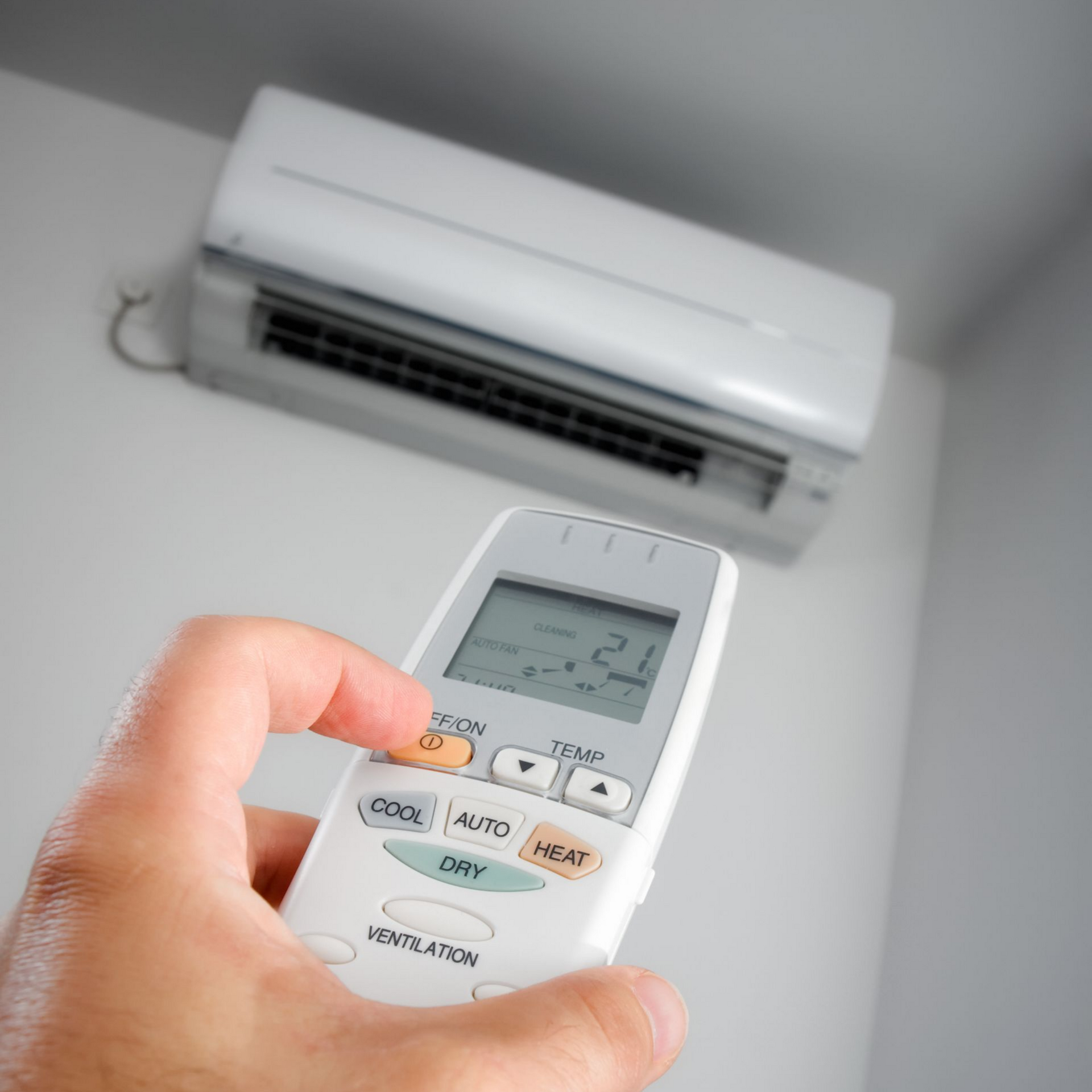 Main tenant la télécommande du climatiseur, pointée vers l'unité murale. Unité blanche avec une grille noire, adossée à un mur blanc.