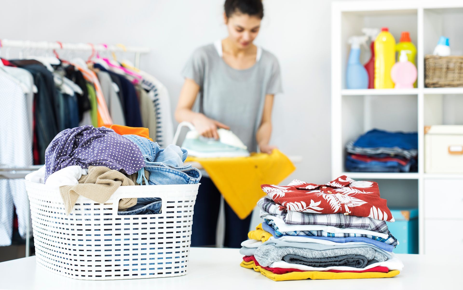 Dans une pièce bien rangée, une personne repasse une chemise jaune derrière un panier à linge et une pile de vêtements pliés.