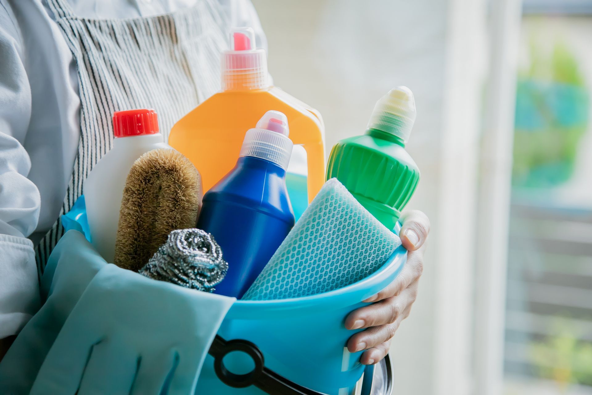 Une personne tenant un seau bleu rempli de divers produits de nettoyage colorés.