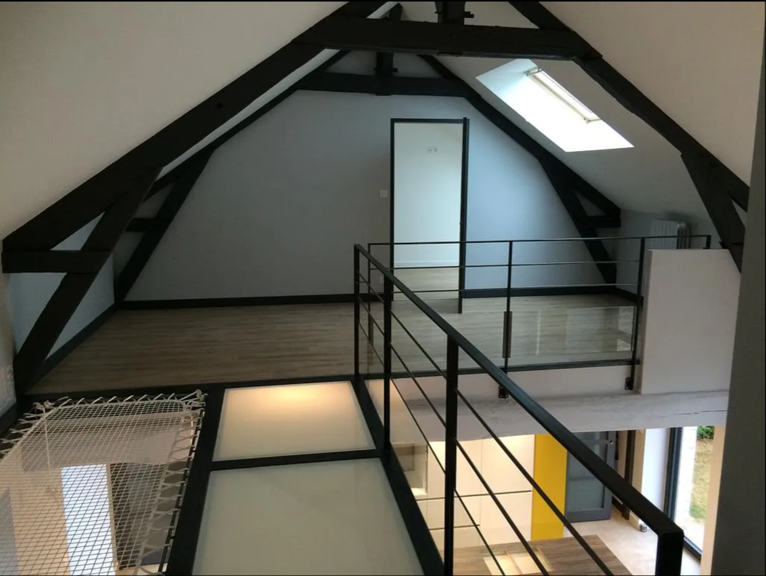 Intérieur de loft avec poutres noires apparentes, puits de lumière, rambarde et porte centrale sur une mezzanine.