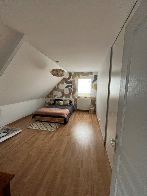 Chambre mansardée étroite avec plafond en pente, lit, tapis à motifs, parquet et une fenêtre lumineuse au fond.