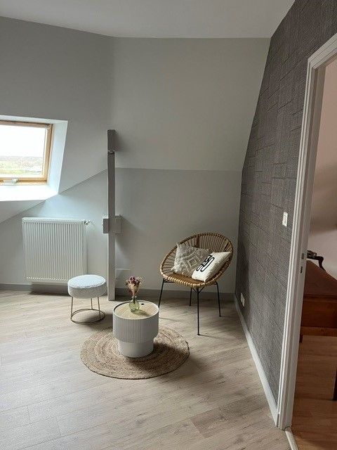 Salon minimaliste avec fauteuil en osier, petite table, tabouret blanc et fenêtre à côté d'un radiateur.