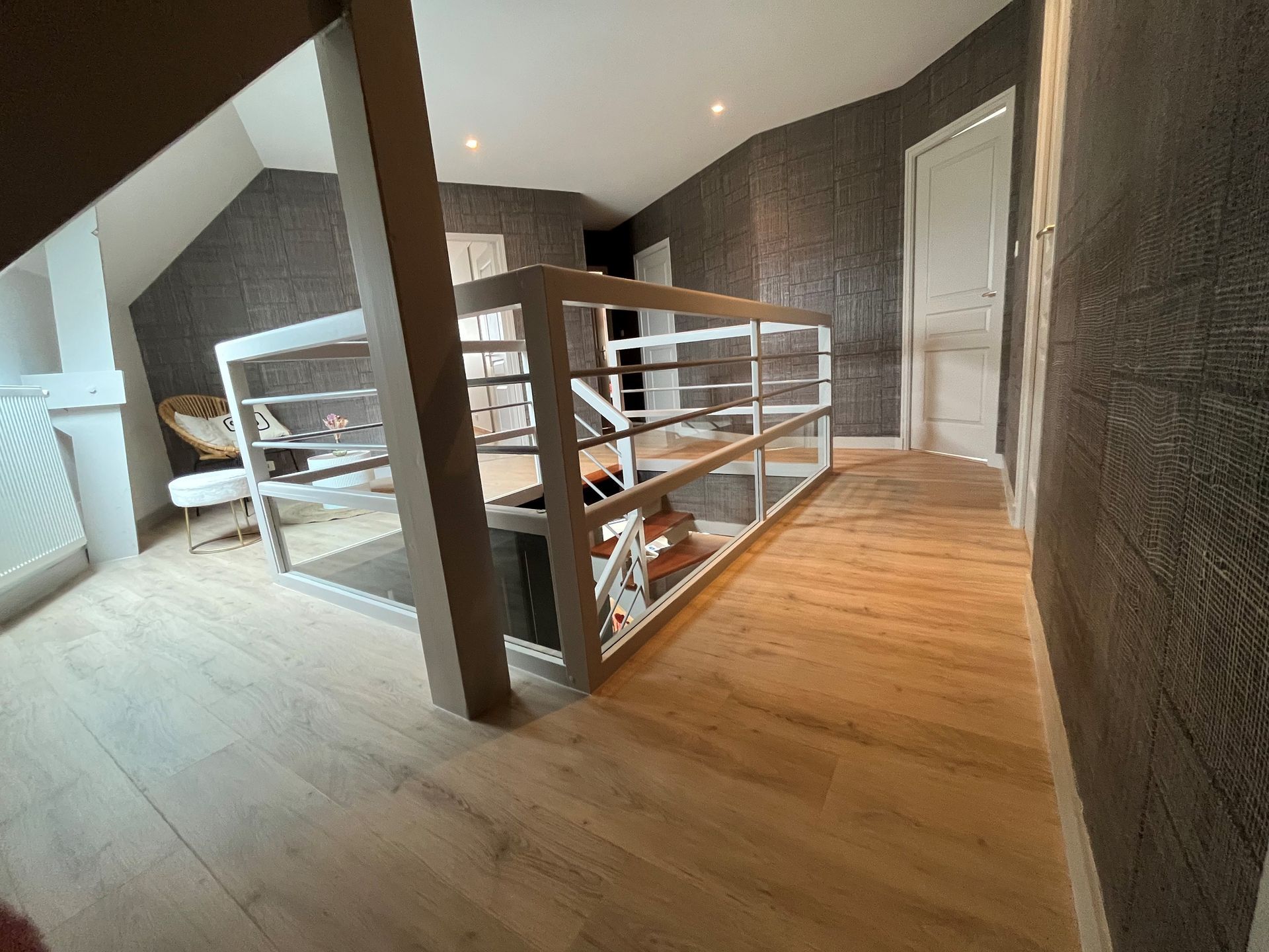 Couloir mansardé avec rampes blanches, parquet et éclairage indirect au plafond.