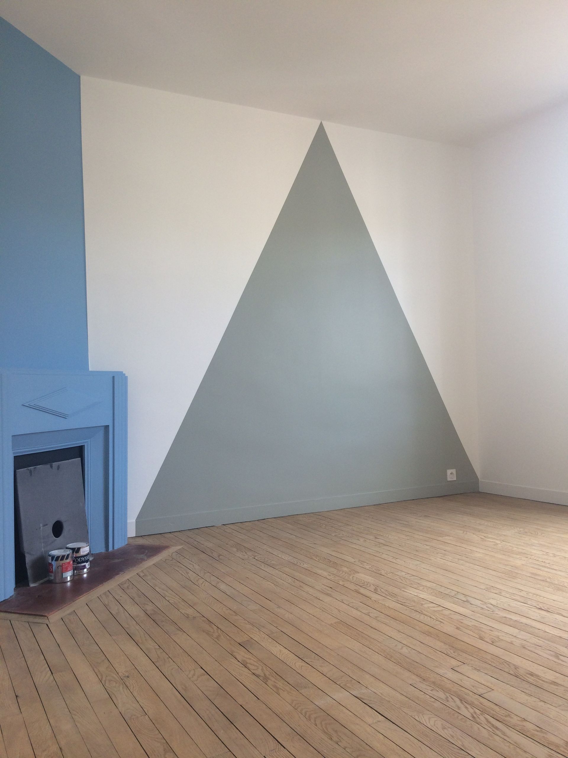 Pièce vide avec cheminée bleu clair et grande fresque murale triangulaire grise sur un mur blanc, parquet