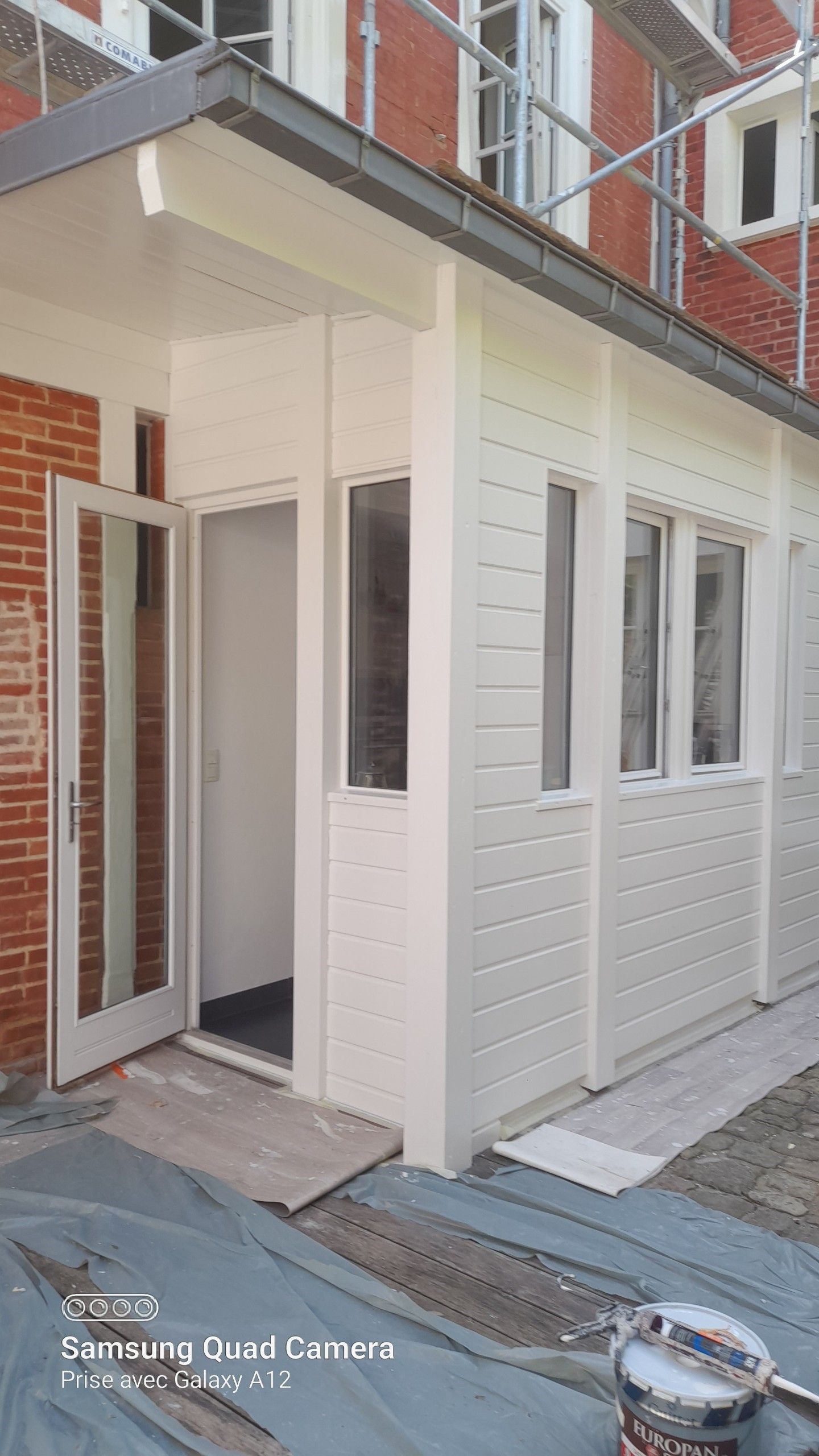 Véranda blanche fermée avec fenêtres et porte ouverte, jouxtant un bâtiment en briques.