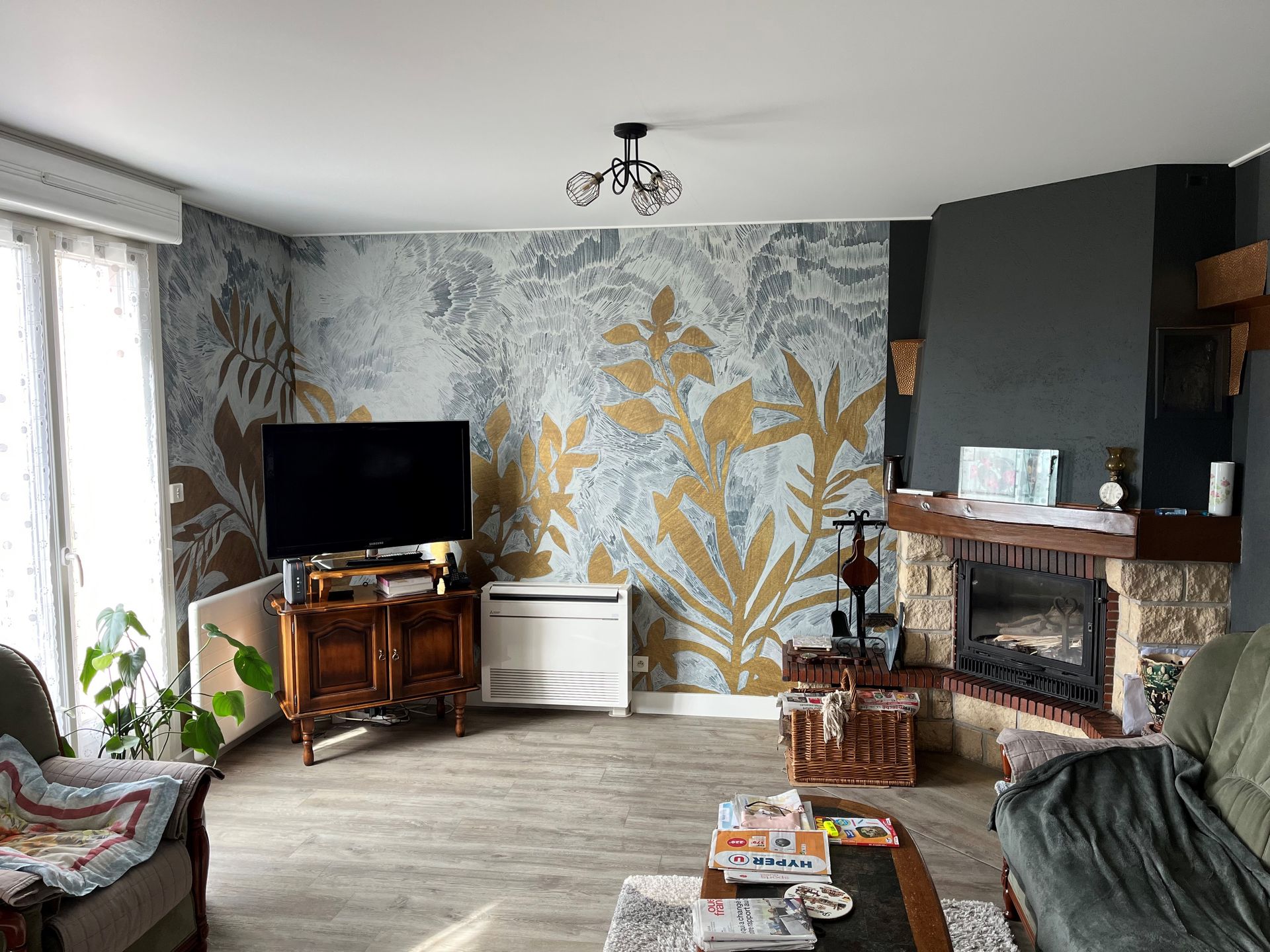 Salon avec mur à motifs floraux, cheminée, télévision et grandes fenêtres avec rideaux à motifs.
