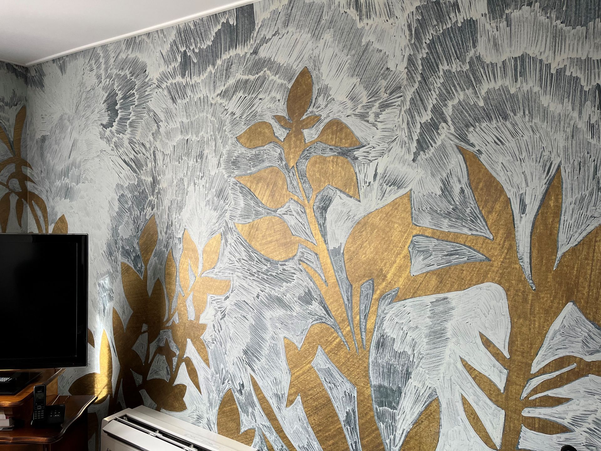 Fresque murale végétale à feuilles dorées sur un mur gris texturé, à côté d'une télévision et d'une petite table.