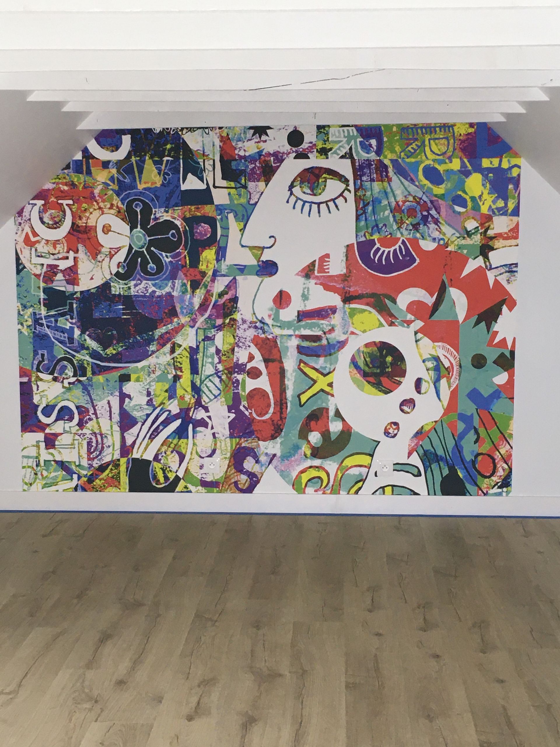 Une fresque murale abstraite et colorée représentant des visages et des motifs orne le mur d'une galerie au-dessus d'un parquet clair.