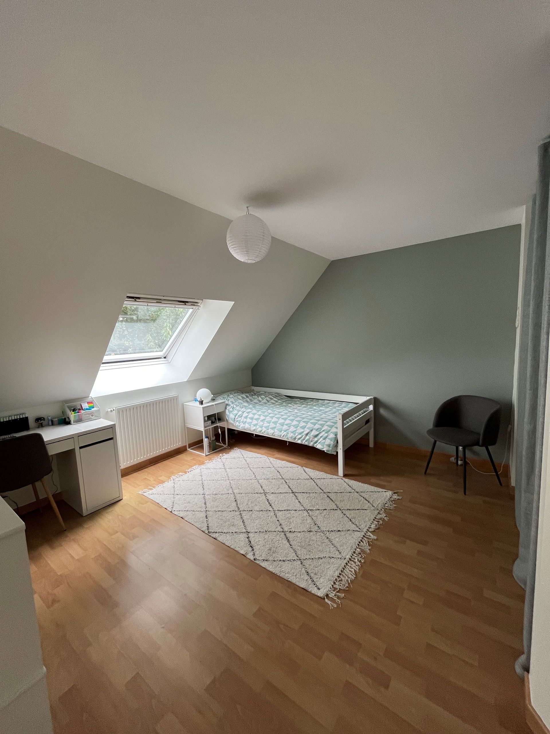 Chambre mansardée épurée avec plafond mansardé, lit, bureau, chaise, tapis et fenêtre de toit.