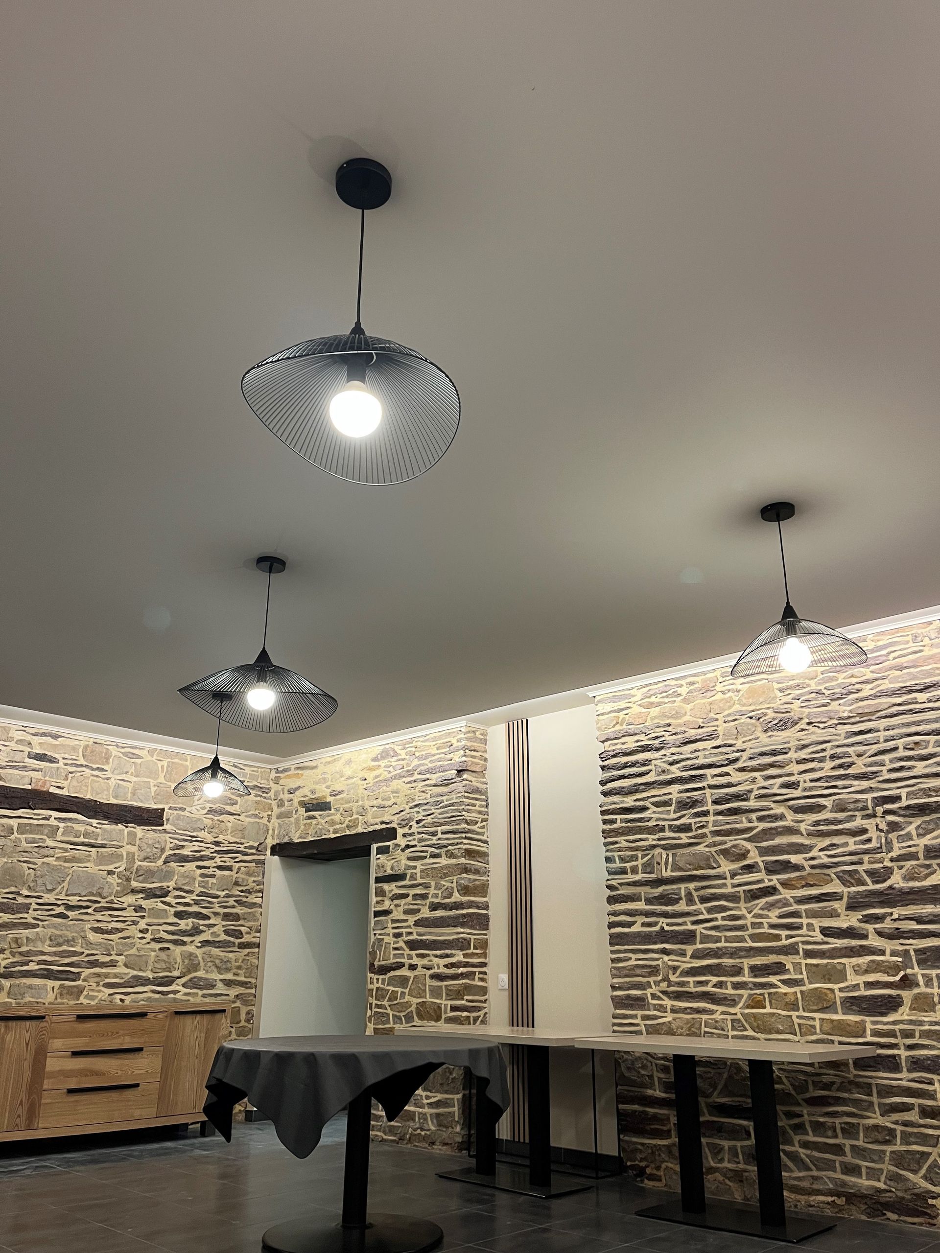 Salle à manger aux murs de pierre, avec luminaires suspendus, tables en bois et une table recouverte d'une nappe.