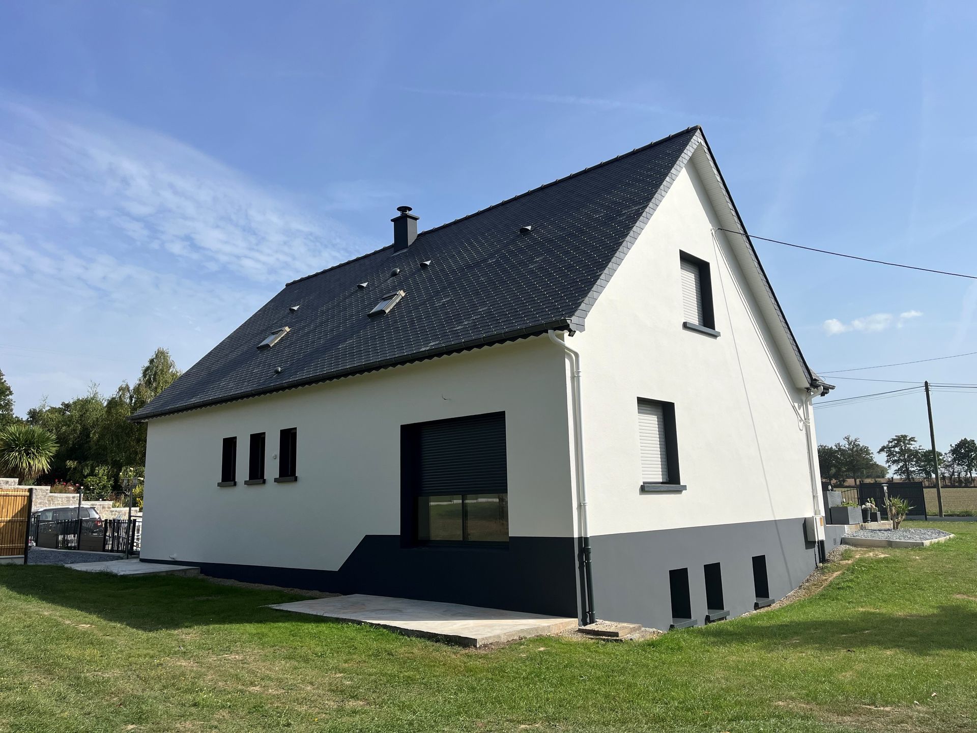 Maison blanche au toit et aux boiseries sombres, vue de côté dans une cour herbeuse sous un ciel bleu.