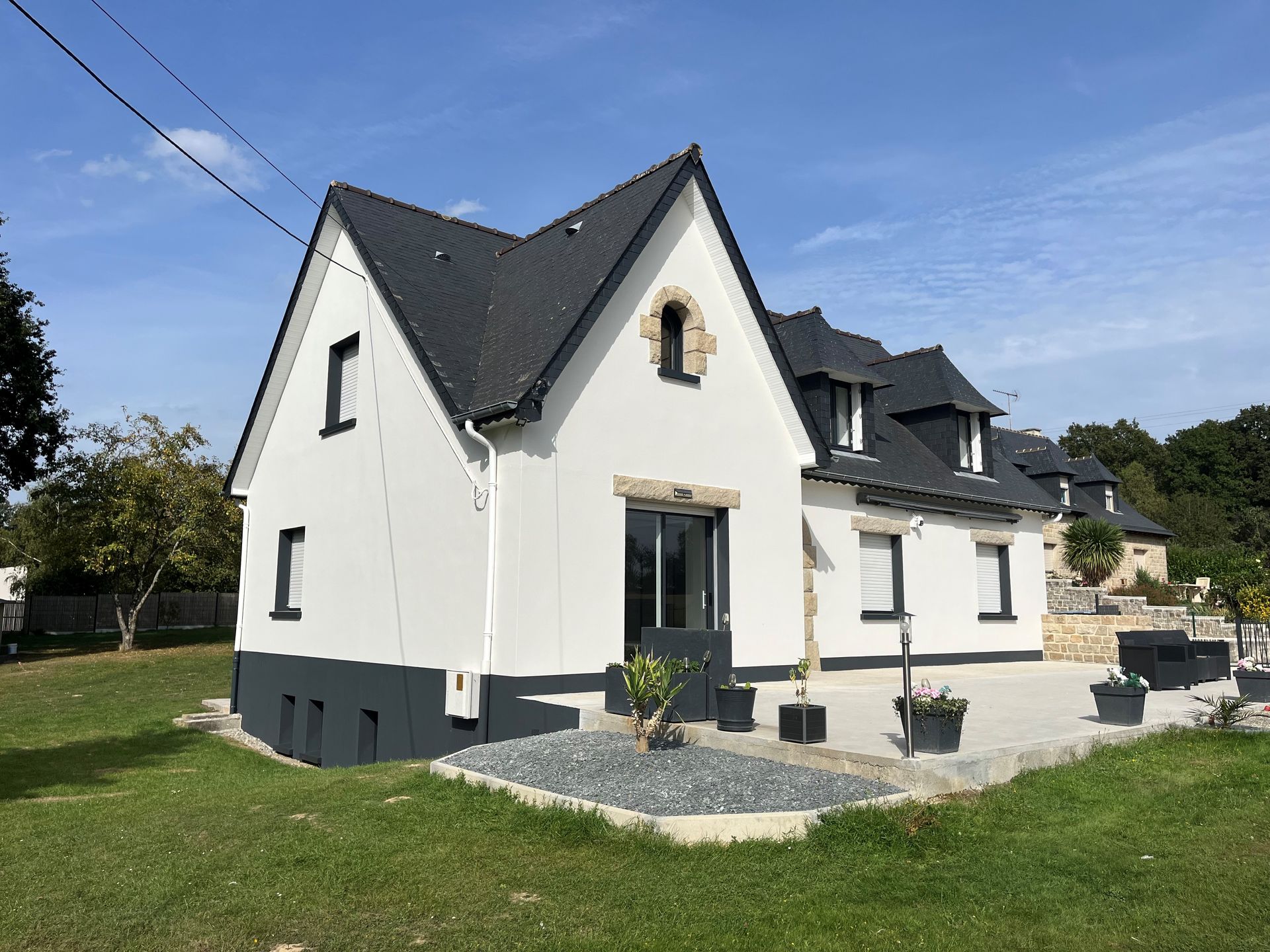 Maison blanche au toit sombre et aux fondations grises, sur un terrain herbeux sous un ciel bleu.