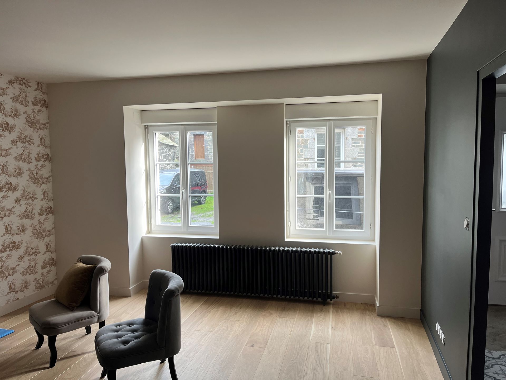 Pièce vide avec deux fenêtres, un radiateur noir, un parquet clair et deux chaises près d'un mur à motifs.