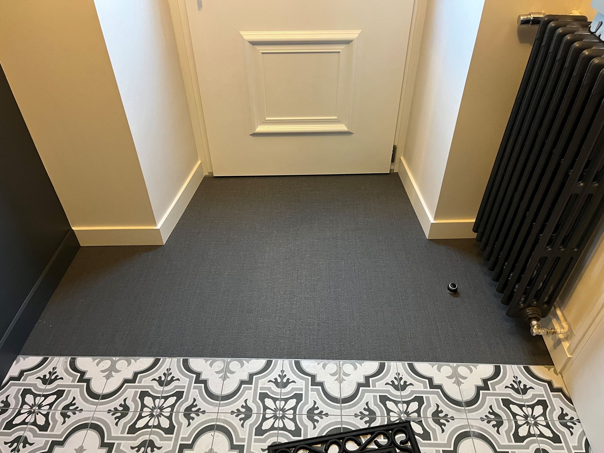 Couloir recouvert de moquette, avec une porte blanche fermée et un radiateur noir sur la droite.