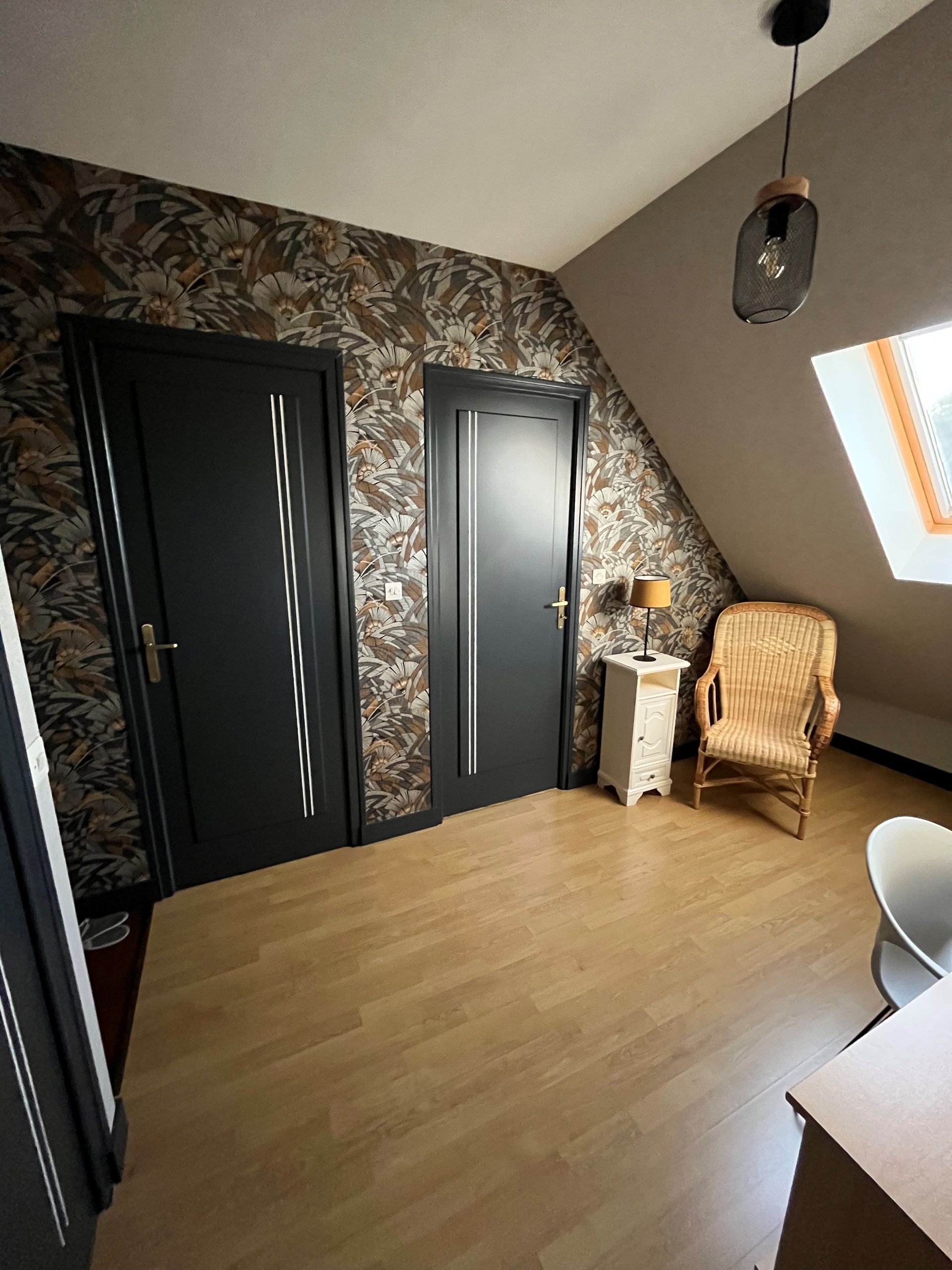 Chambre mansardée moderne avec papier peint fleuri, deux portes sombres, fauteuil en osier et fenêtre de toit.