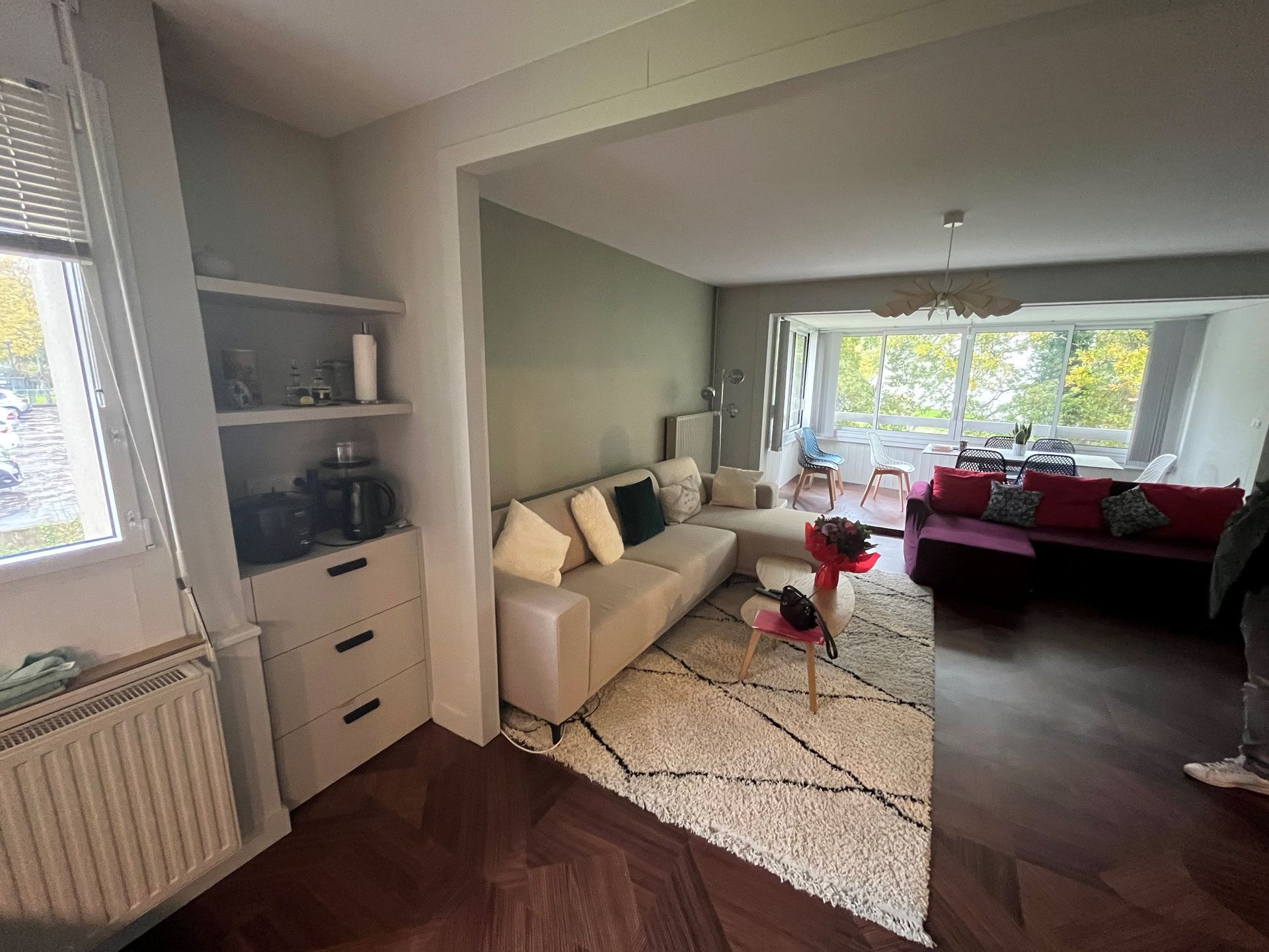 Salon confortable avec canapé beige, tapis à motifs, fauteuils rouges et grandes fenêtres donnant sur les arbres