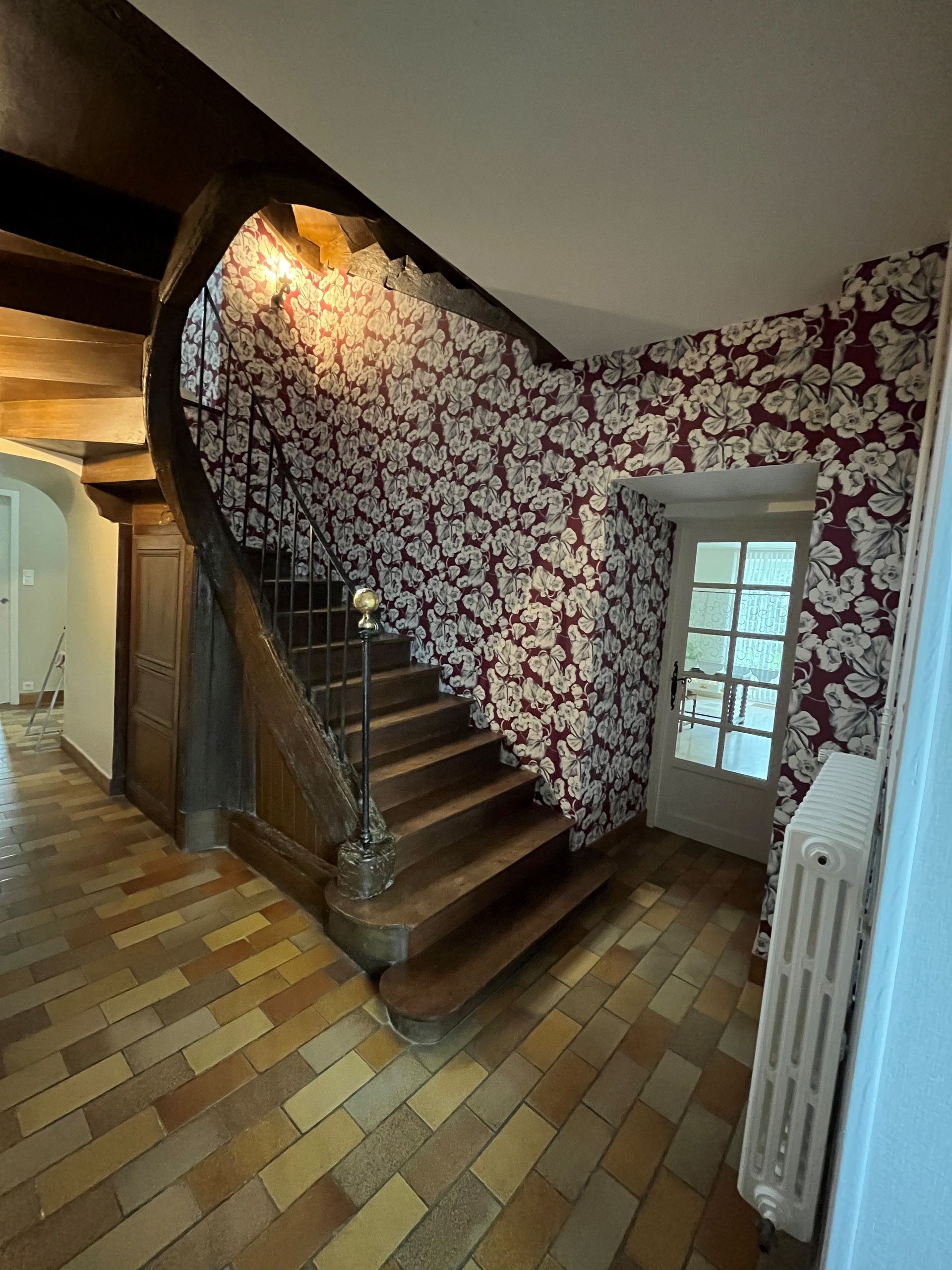 Escalier dans un couloir étroit avec papier peint à fleurs, sol carrelé à motifs et radiateur blanc