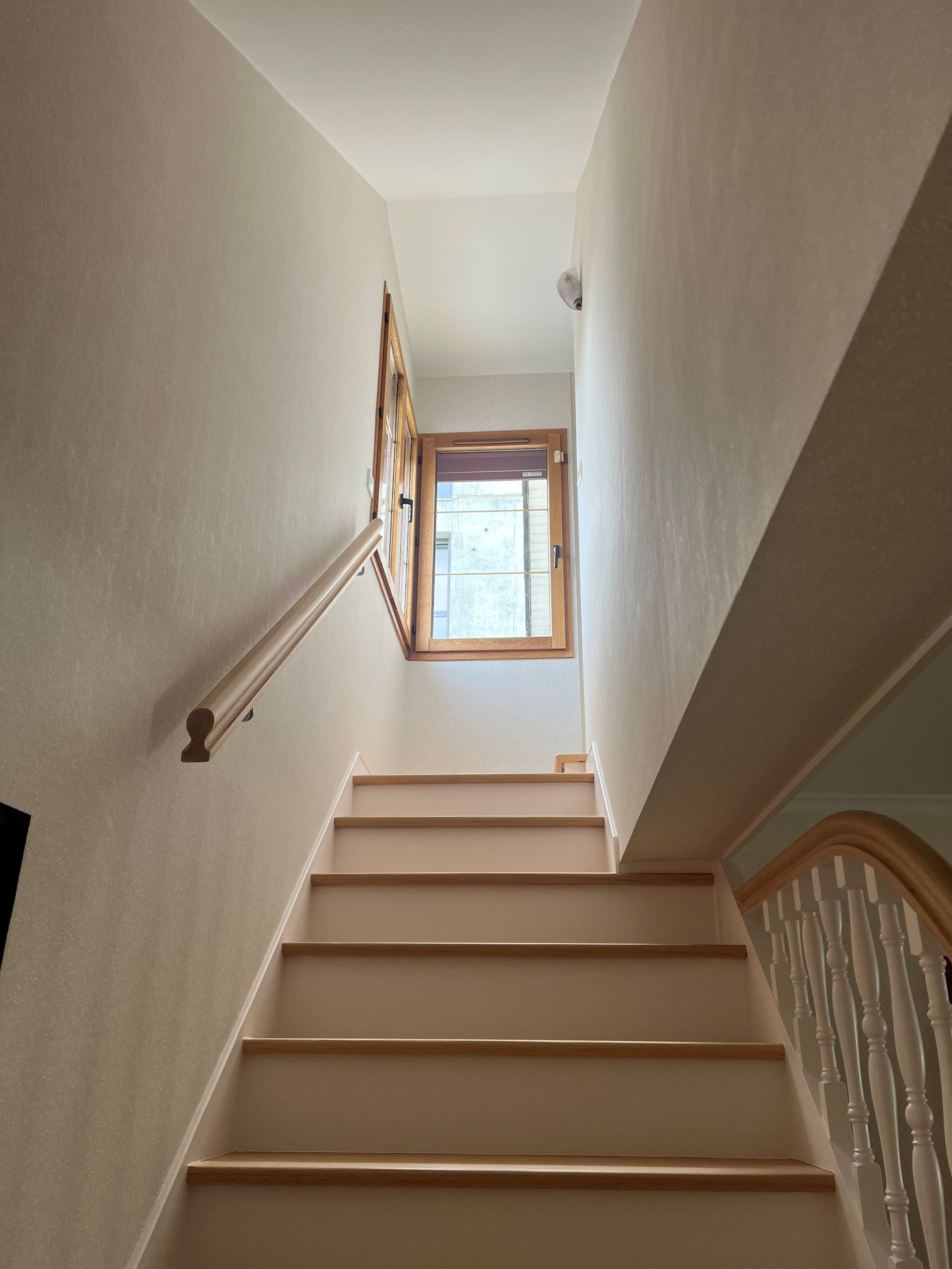 Escalier intérieur étroit aux murs beiges, marches en bois et fenêtre en haut.