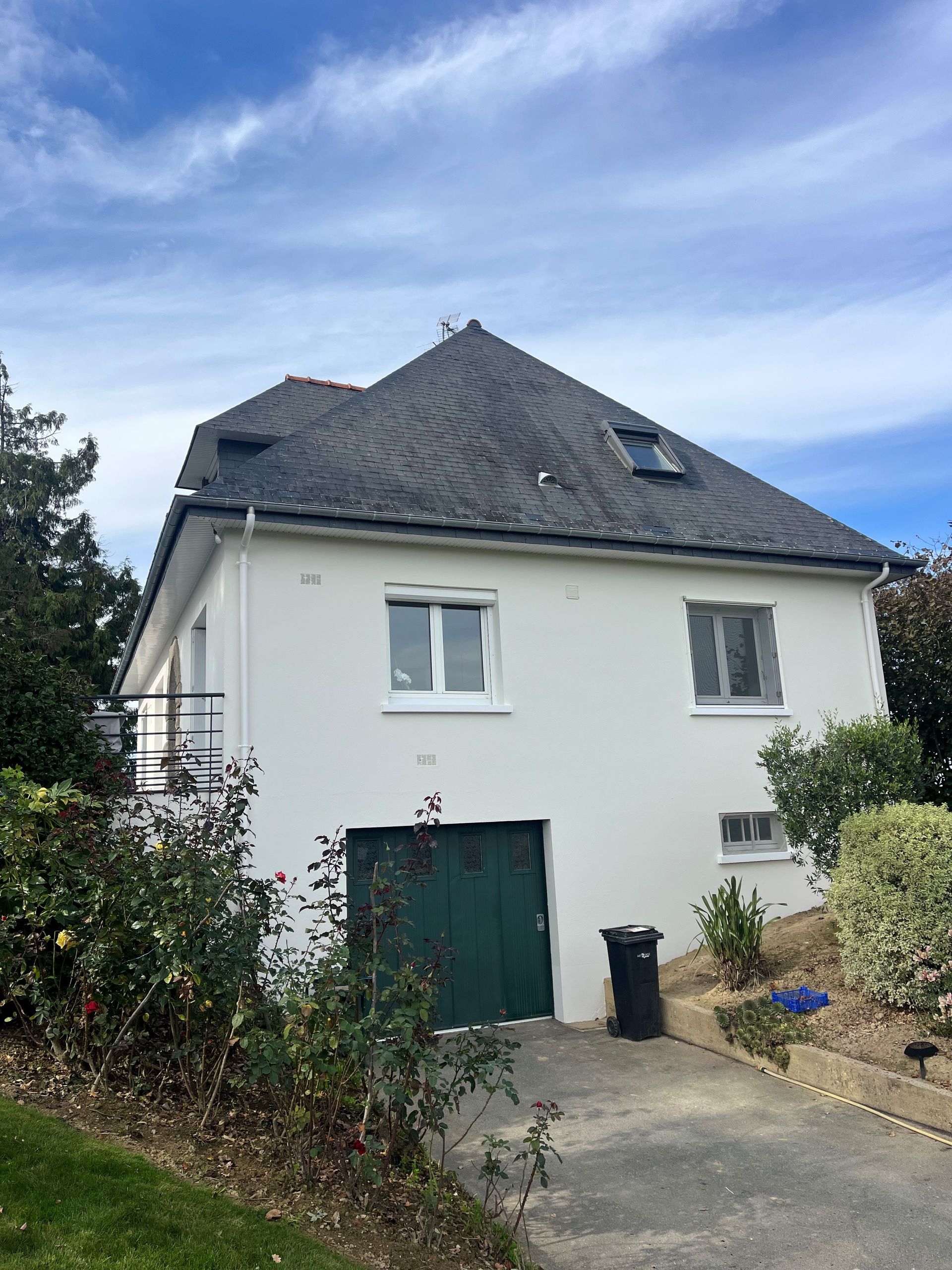 Maison blanche à deux étages avec toit foncé, porte de garage verte et allée sous un ciel nuageux