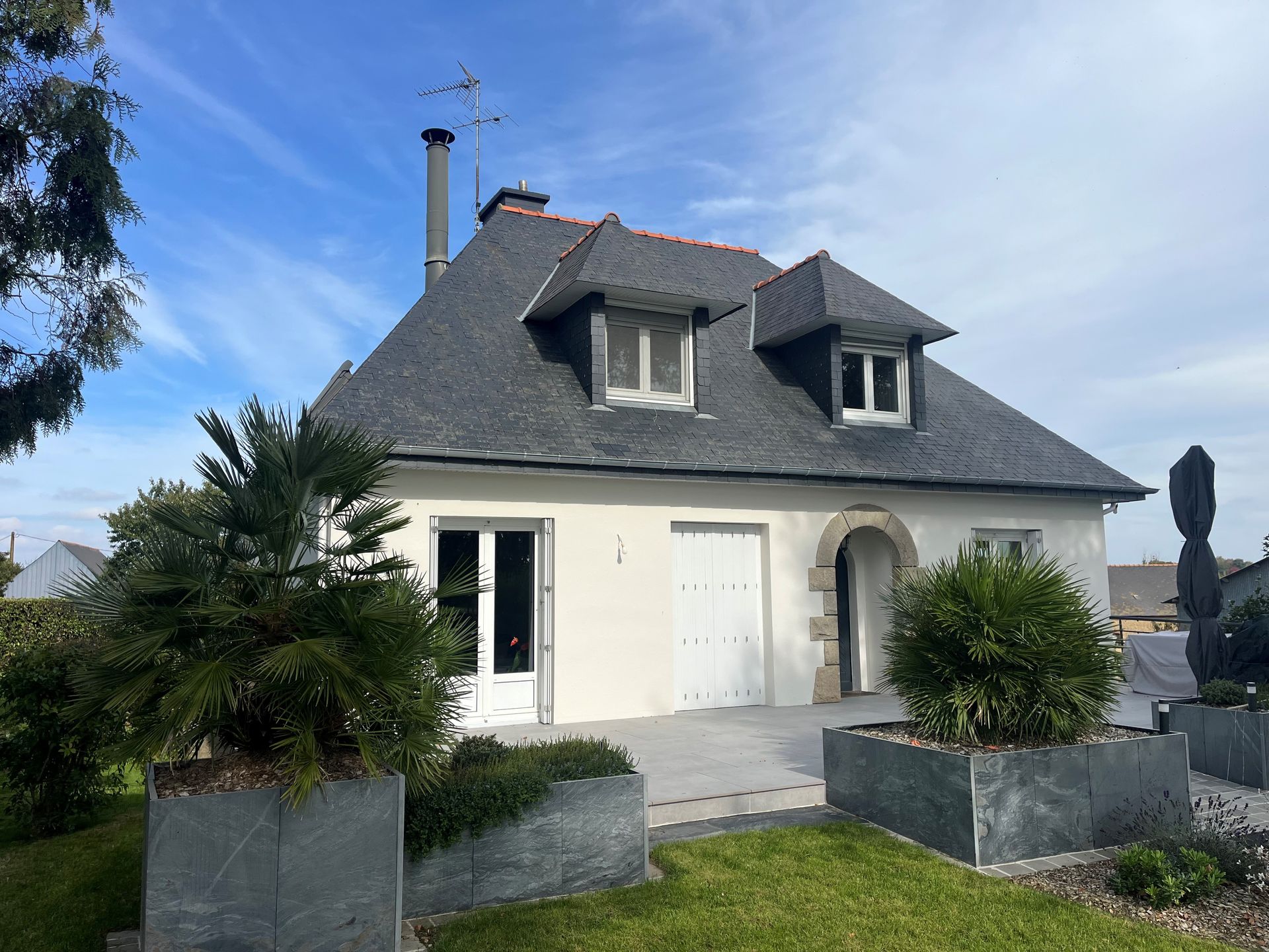 Maison blanche au toit sombre, entrée cintrée, lucarnes et plantes en pot par une journée ensoleillée