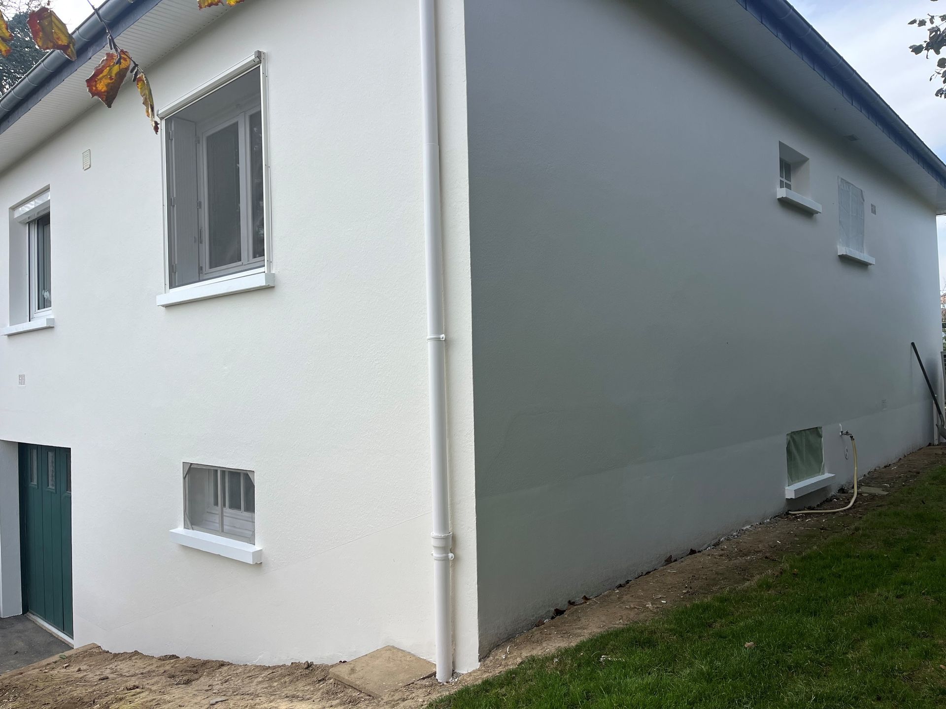 Maison blanche avec un mur latéral gris, des fenêtres, une porte et une pente herbeuse à côté.