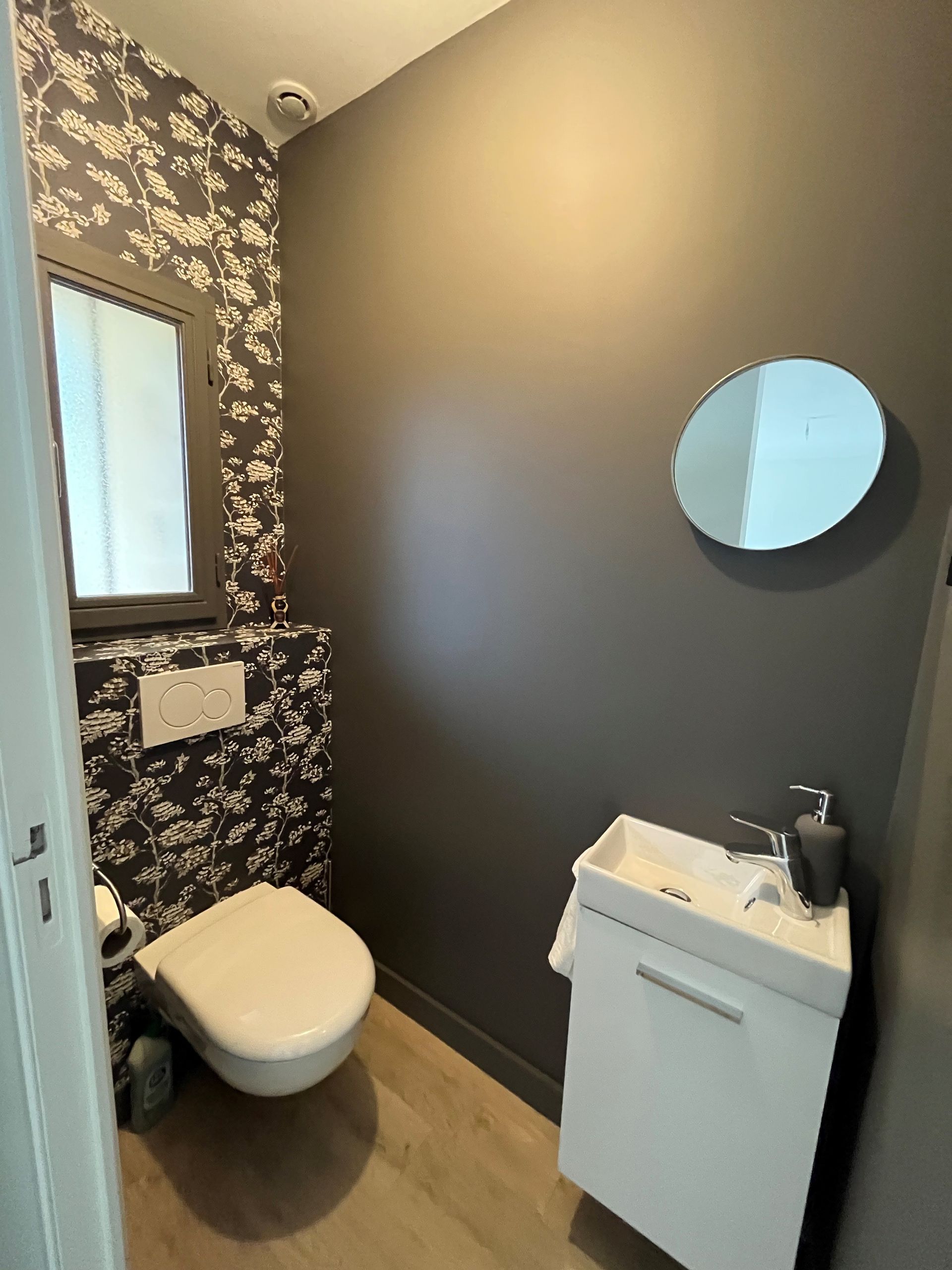 Petite salle de bains avec papier peint à motifs floraux foncés, mur gris, toilettes, meuble-lavabo et miroir rond.