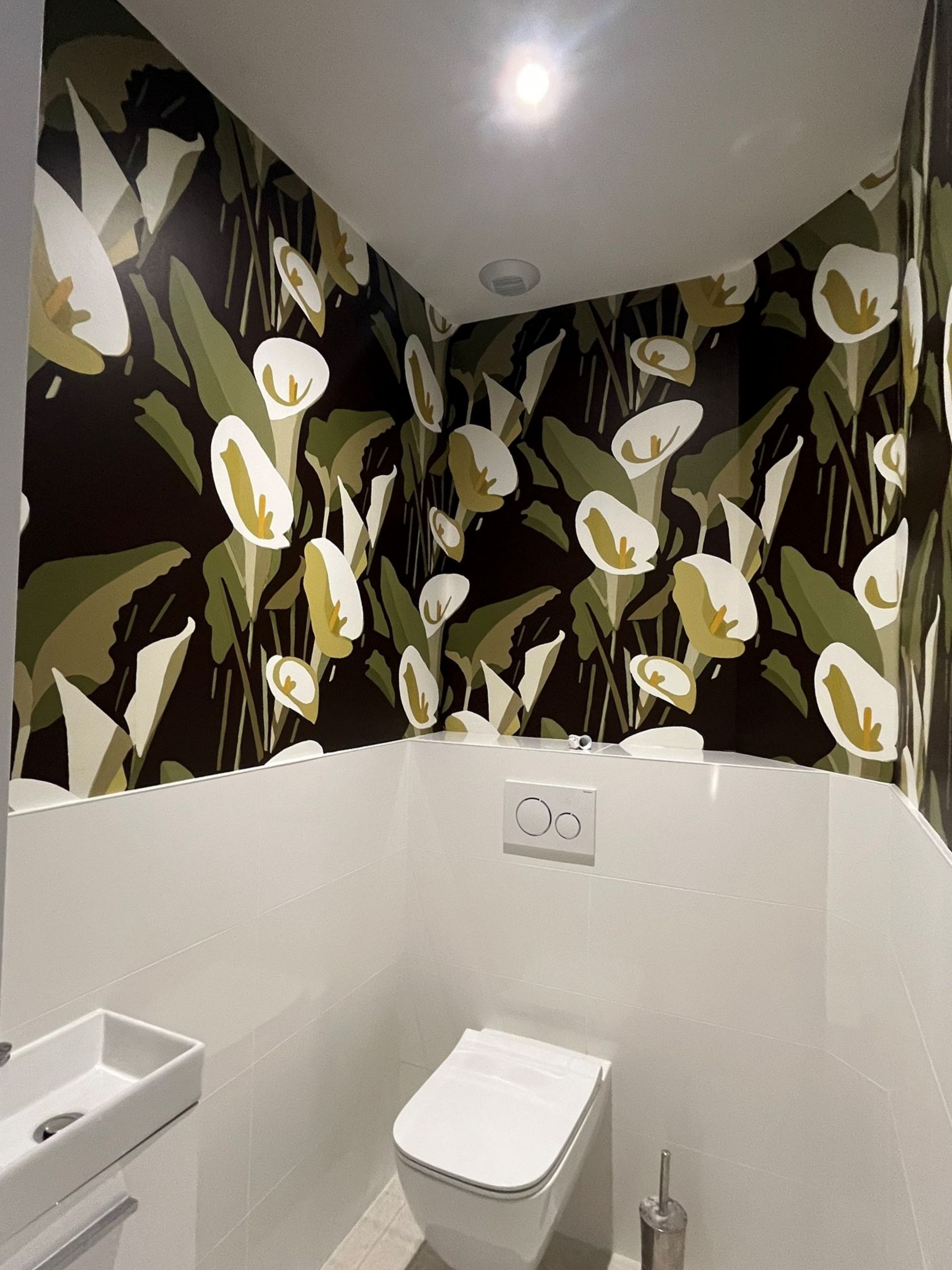 Petite salle de bains avec un papier peint à motifs floraux audacieux au-dessus de carreaux blancs et des toilettes suspendues sous un plafonnier.