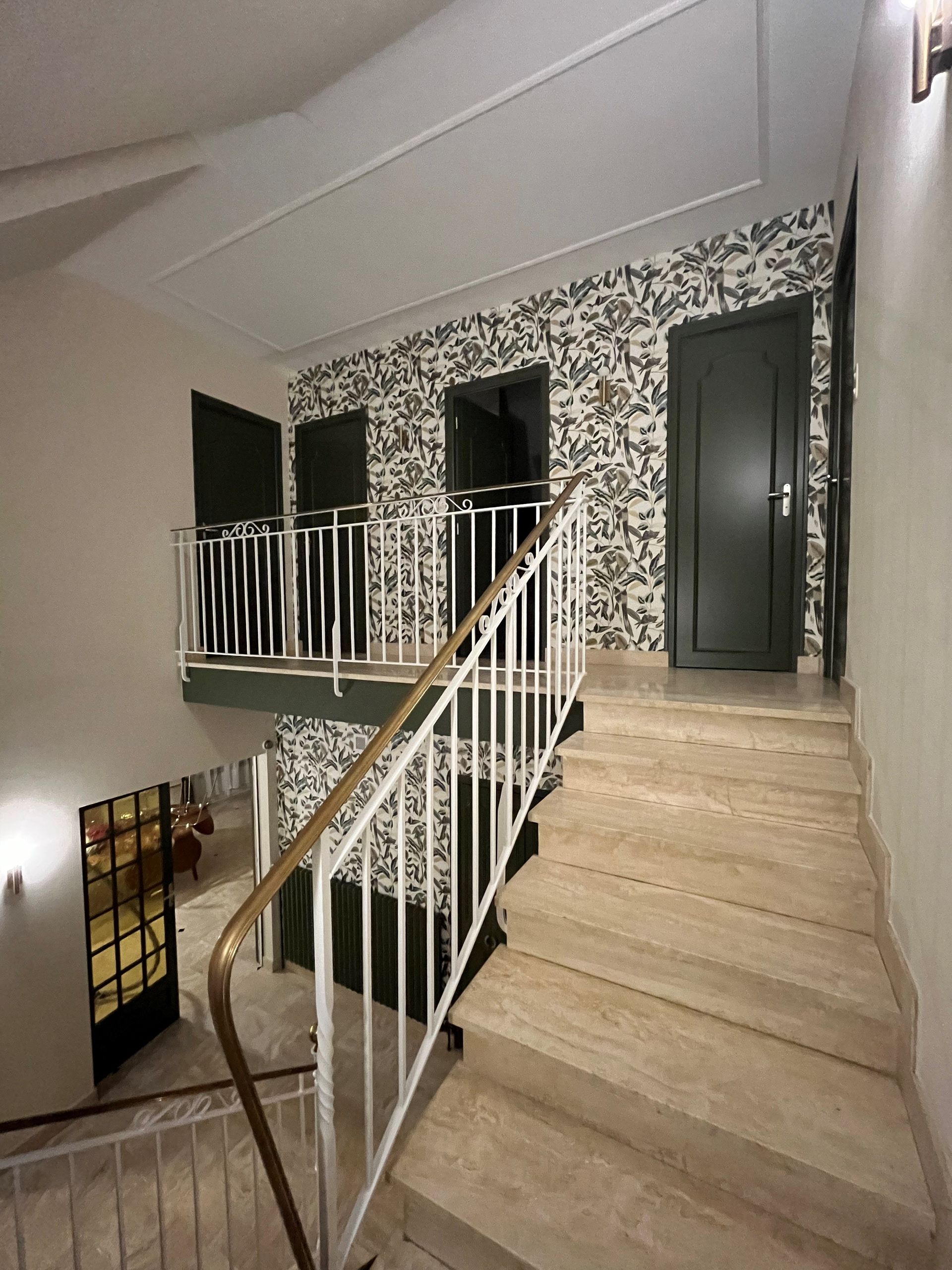 Escalier avec moquette beige et rampe blanche menant à un couloir tapissé de papier peint à motifs floraux noirs et blancs.