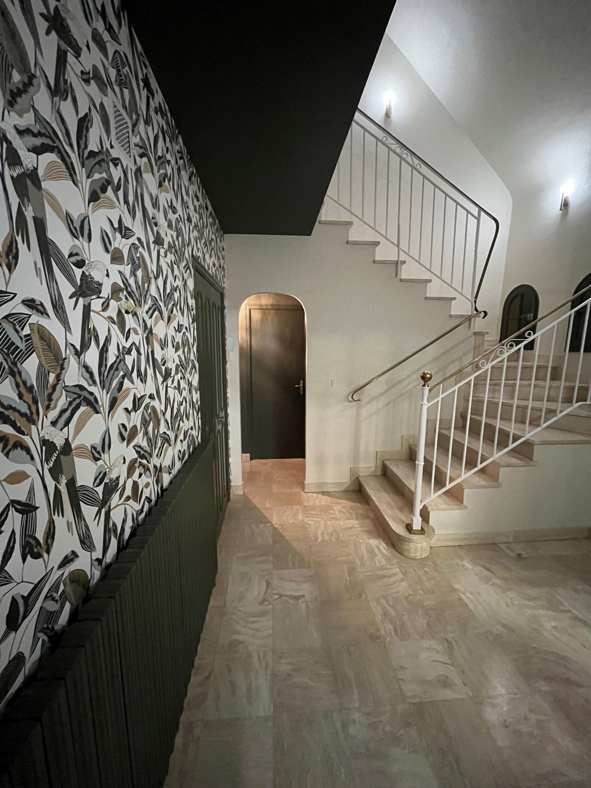 Cage d'escalier vide avec mur à motifs noirs et blancs, sol carrelé beige et escalier blanc sous une lumière vive