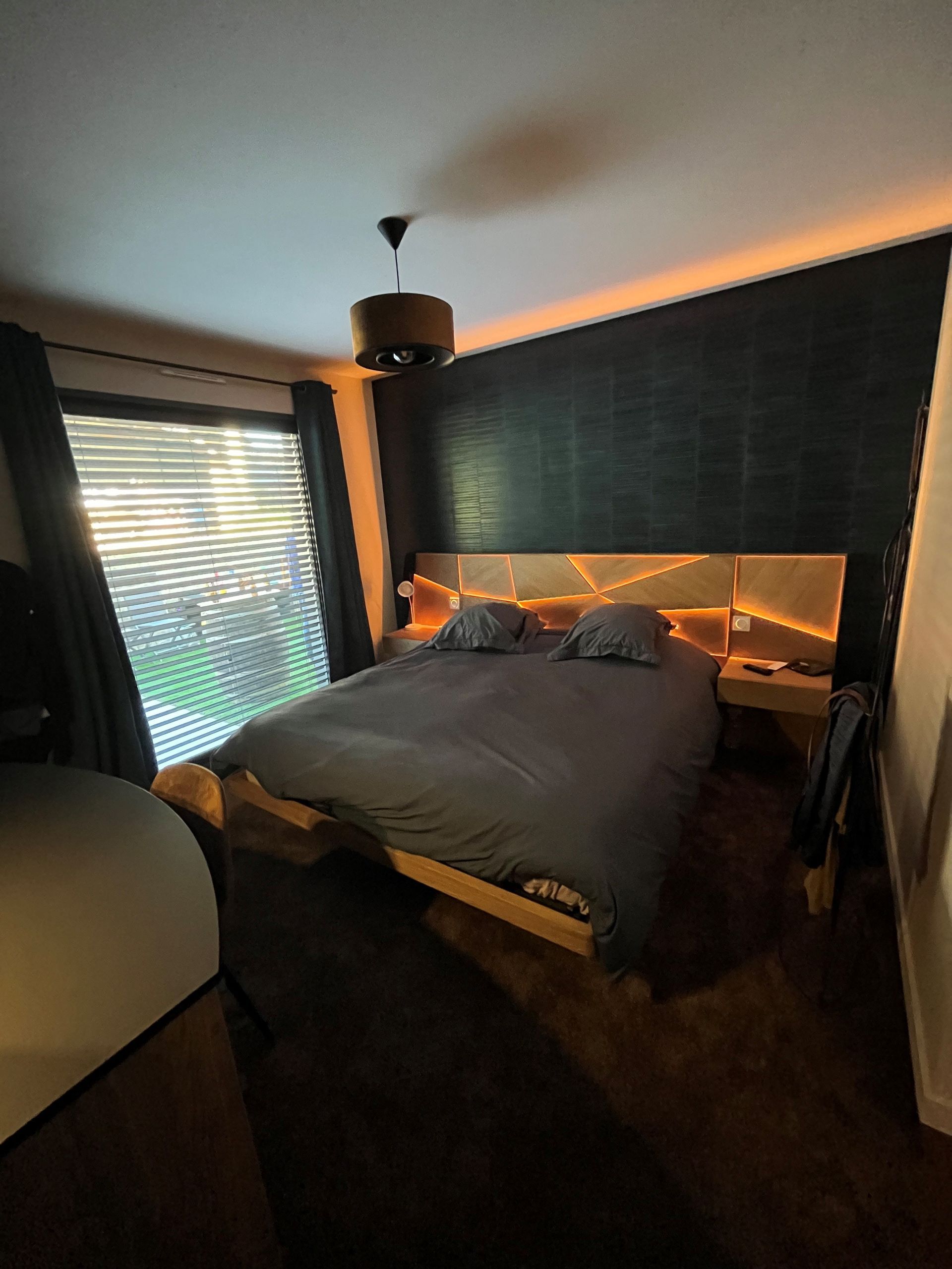 Chambre confortable avec un lit, un éclairage LED chaleureux au niveau de la tête de lit, des murs sombres et une fenêtre avec stores.