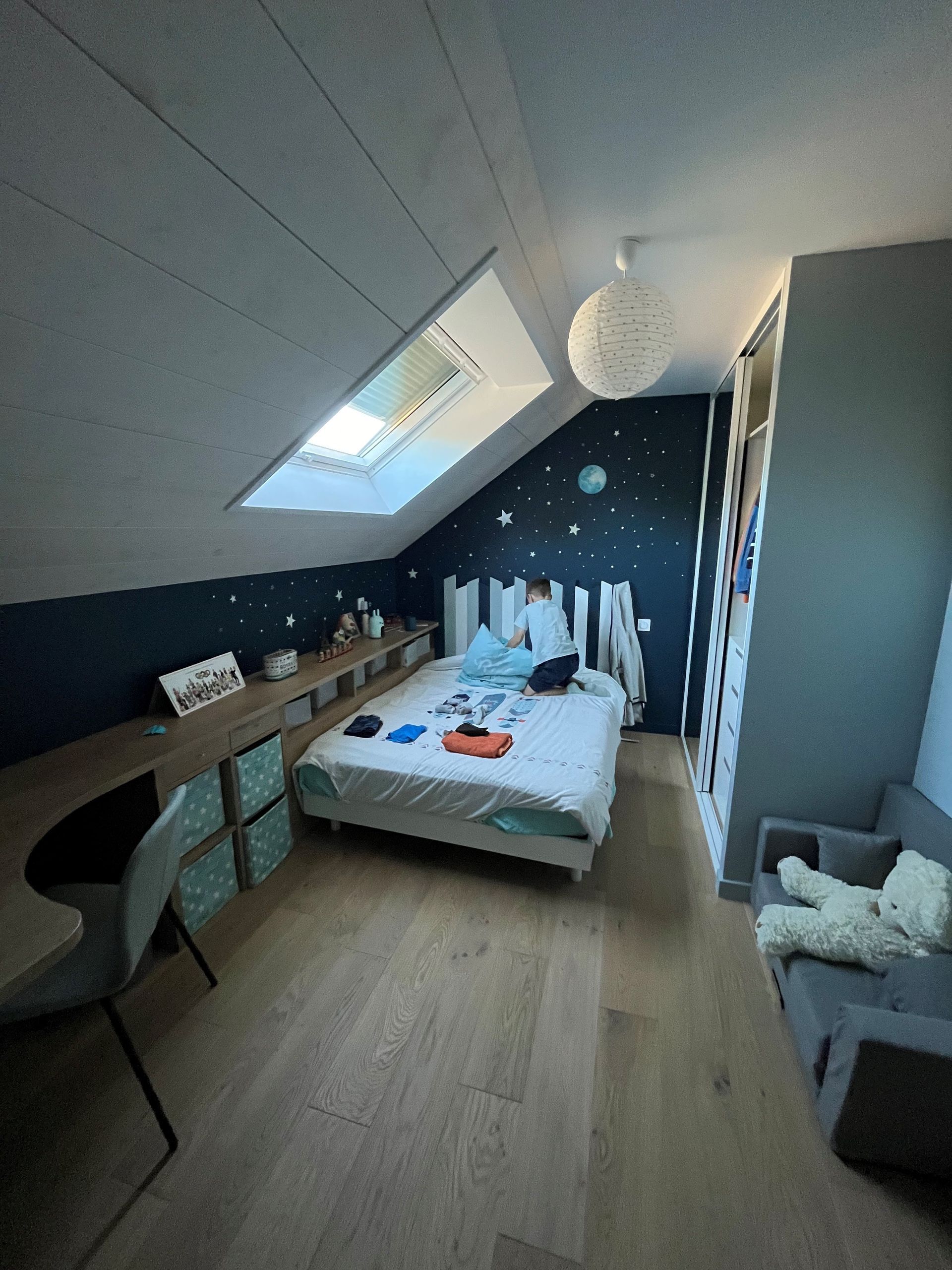 Petite chambre mansardée avec puits de lumière, lit bas, mur d'accent étoilé foncé et parquet clair.