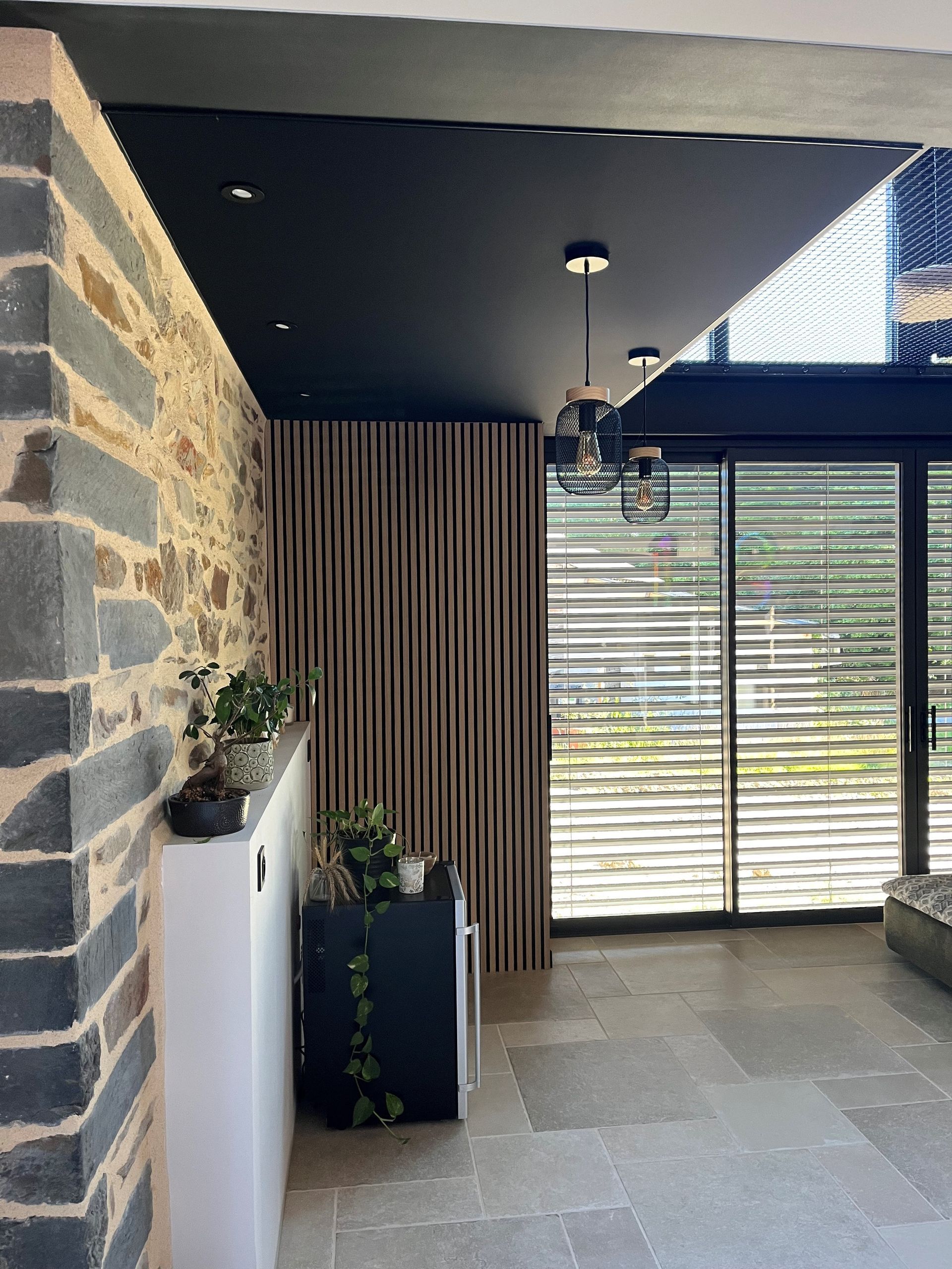 Patio couvert avec mur en pierre, sol en carrelage gris, luminaires suspendus et porte coulissante en verre avec stores à rayures.