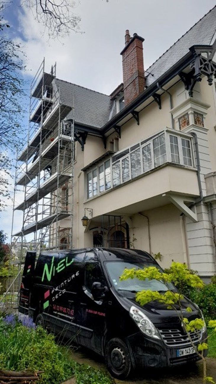 Une camionnette de service noire est garée à côté d'une maison couleur crème, avec des échafaudages sur le côté gauche.