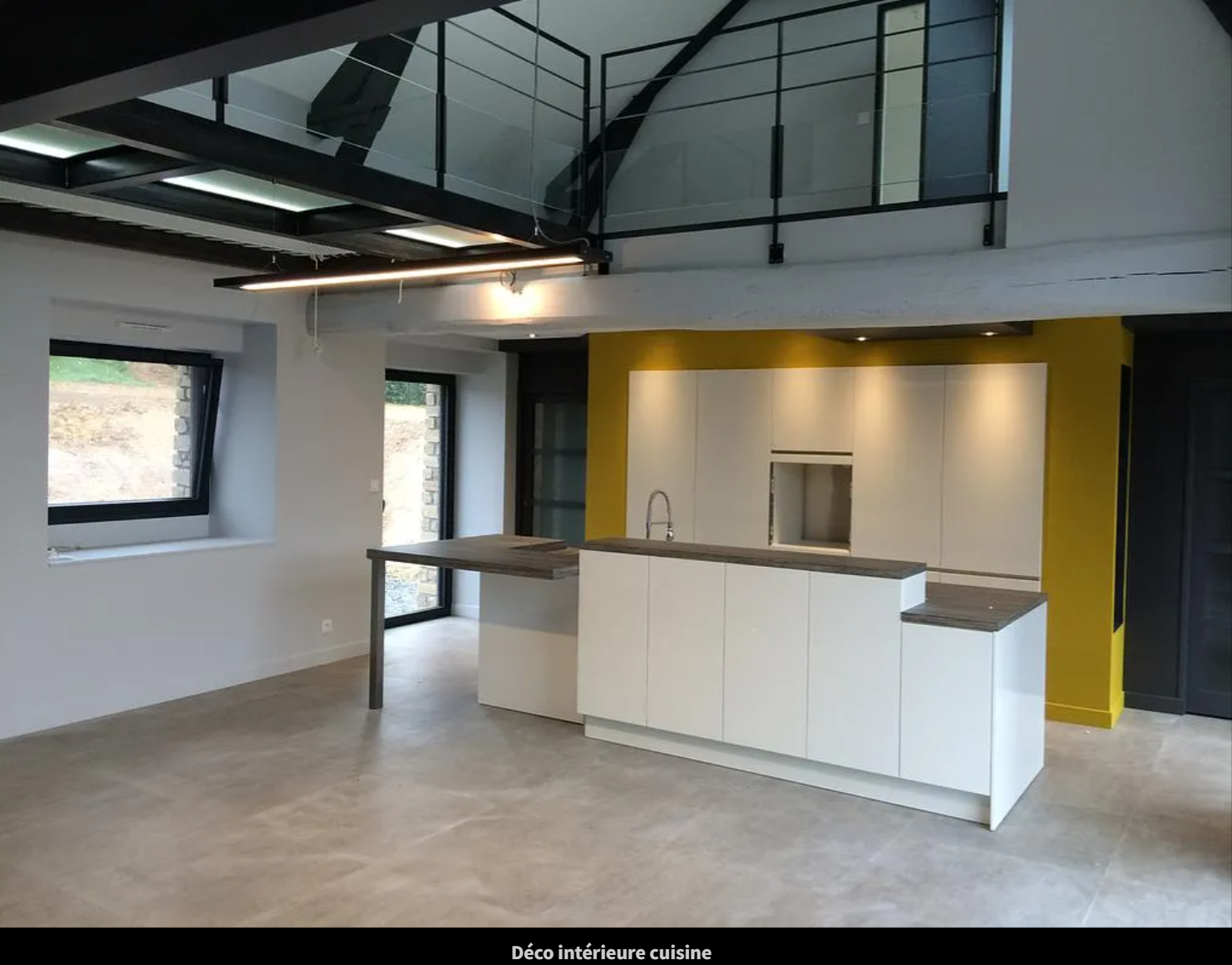 Cuisine moderne ouverte avec îlot central blanc, sol en béton, hauts plafonds et fenêtres de style industriel