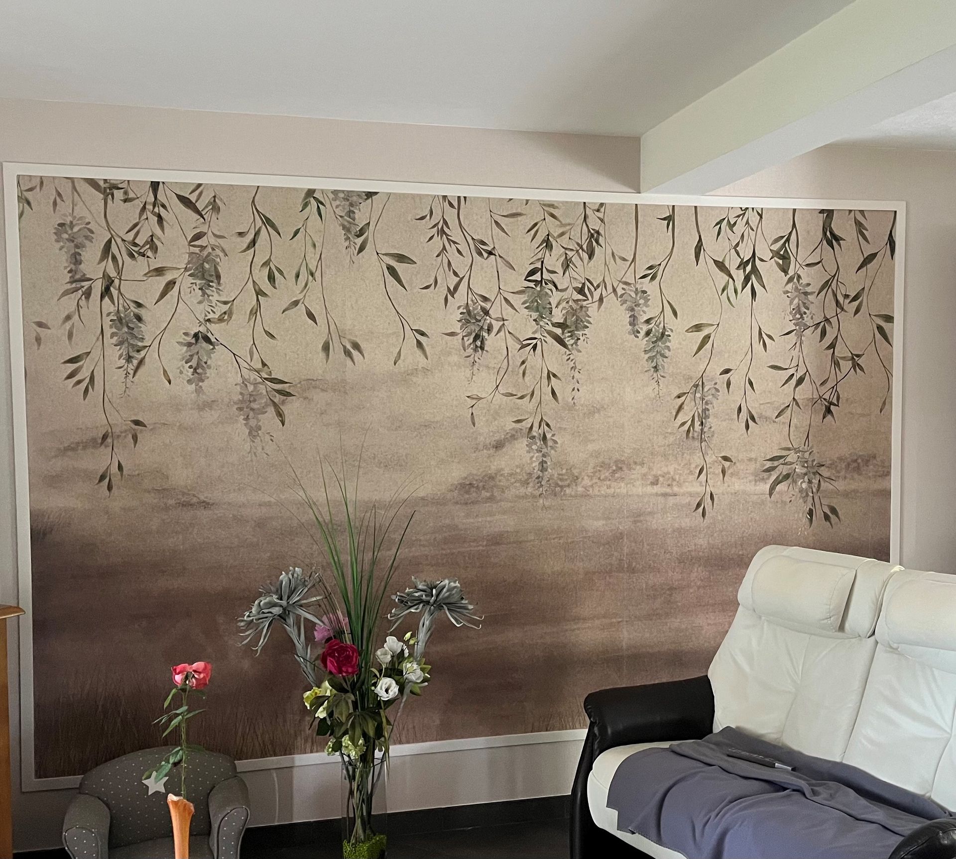 Un salon avec une grande fresque murale représentant des branches suspendues, une chaise blanche et des fleurs dans un vase.