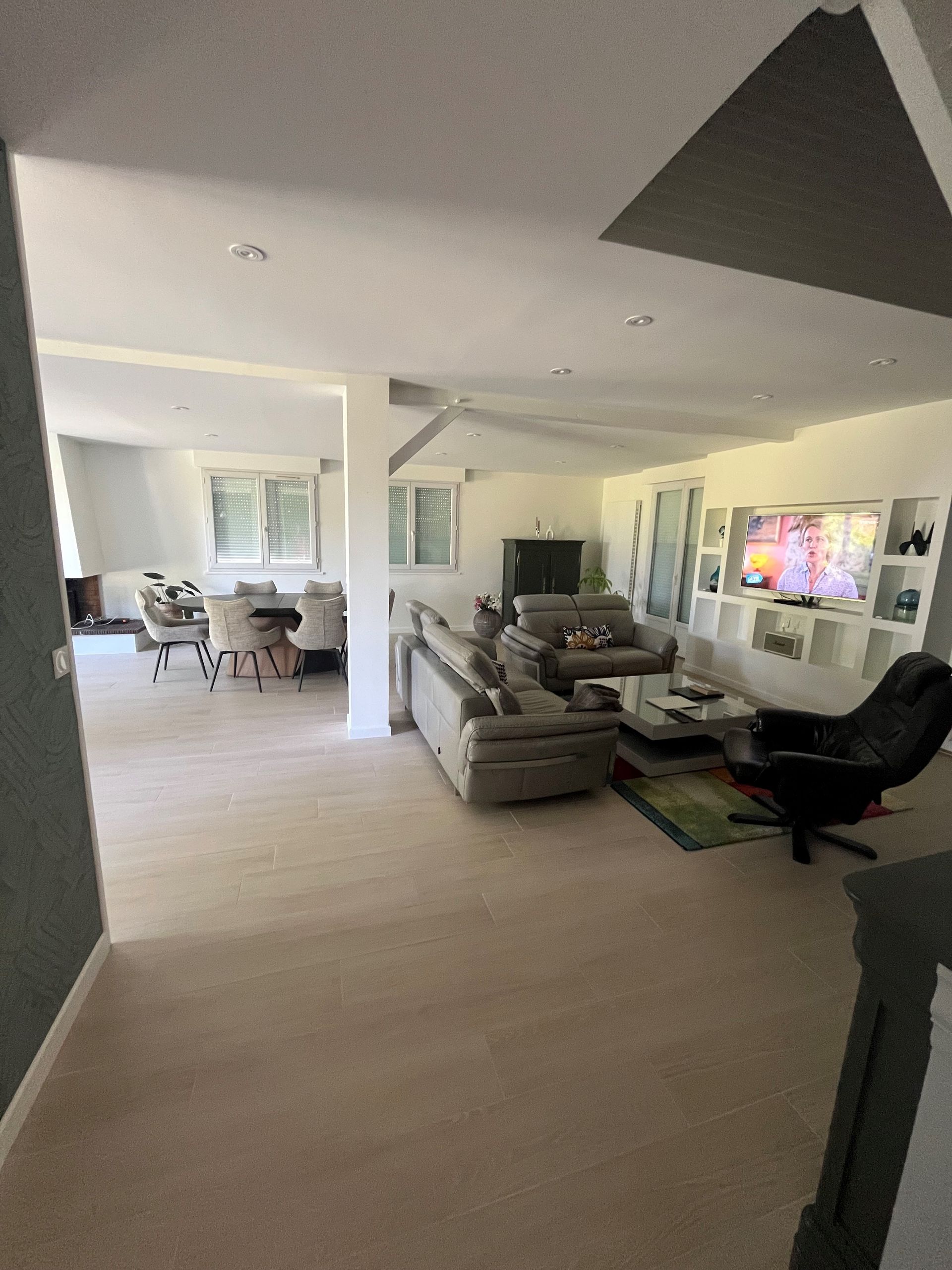 Salon lumineux avec canapé d'angle gris, fauteuil noir, télévision murale et coin repas ouvert attenant.