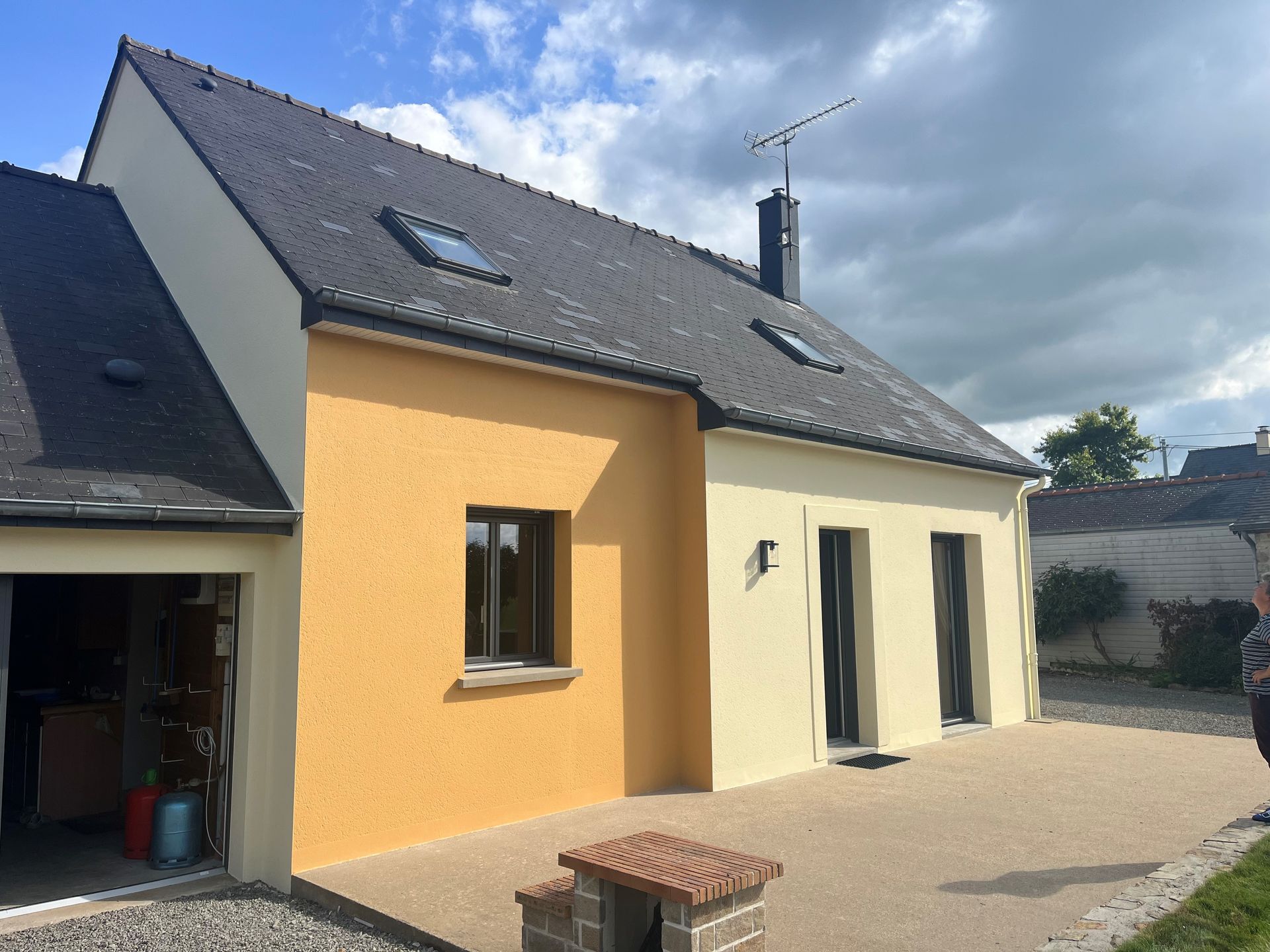 Maison jaune au toit gris, puits de lumière et garage ouvert sous un ciel nuageux