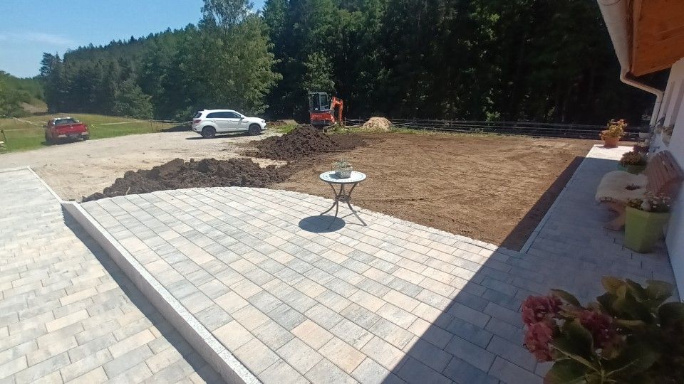 Terrasse mit Pflastersteinen, unbewachsenem Erdreich und kleinem Tisch; im Hintergrund ein weißes Auto und Bäume.