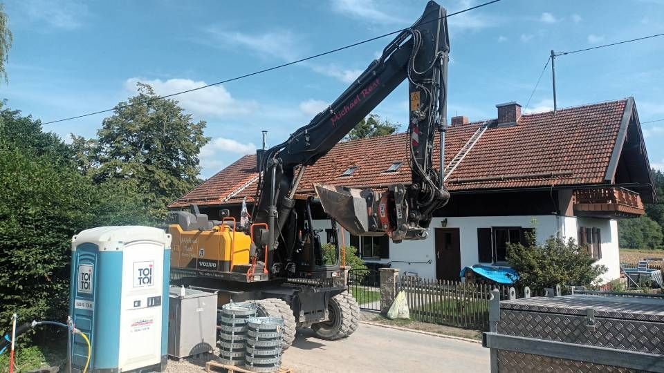 Ein Bagger reißt ein Gebäude ab; im Hintergrund sind eine mobile Toilette und ein Wohnhaus zu sehen.