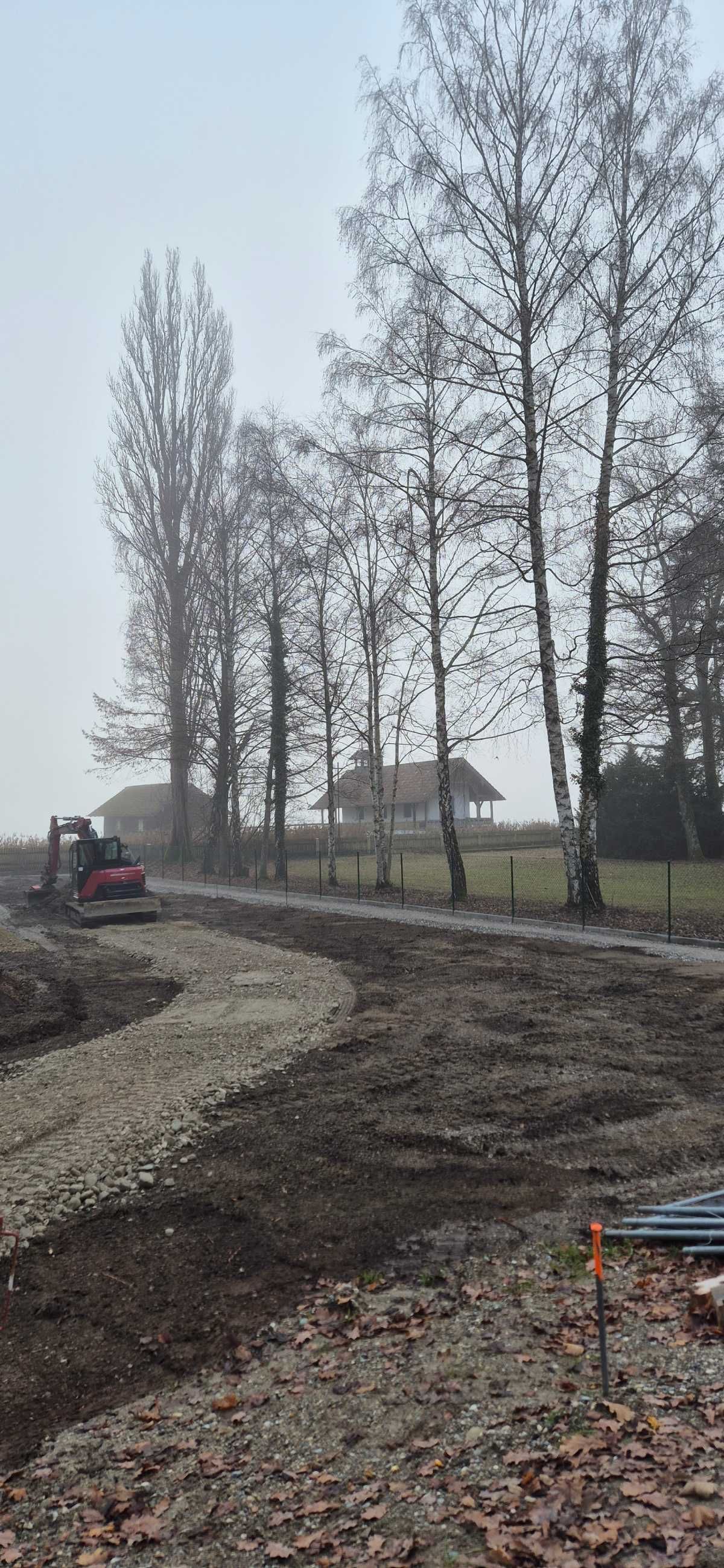 Ein gewundener Schotterweg wird mit einem kleinen Bagger angelegt. Im Hintergrund sind kahle Bäume und ein Haus zu sehen.
